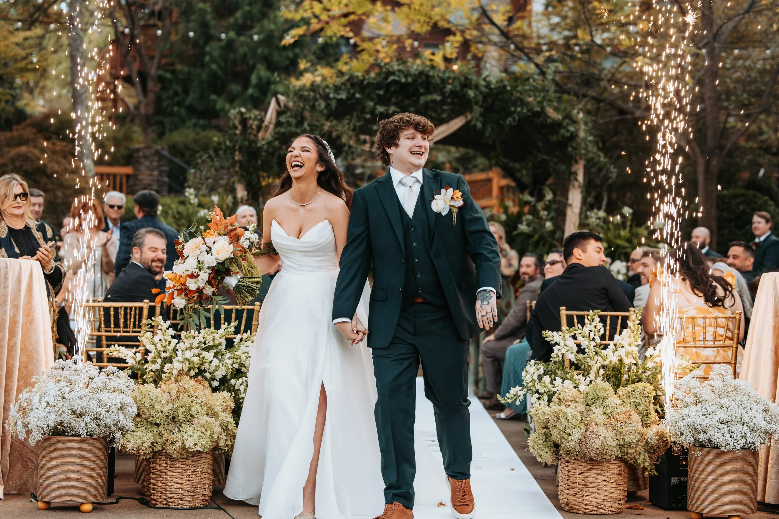 Bride and groom walking hand in hand at their outdoor wedding reception, surrounded by guests, floral arrangements, and sparklers, with autumn trees in the background.