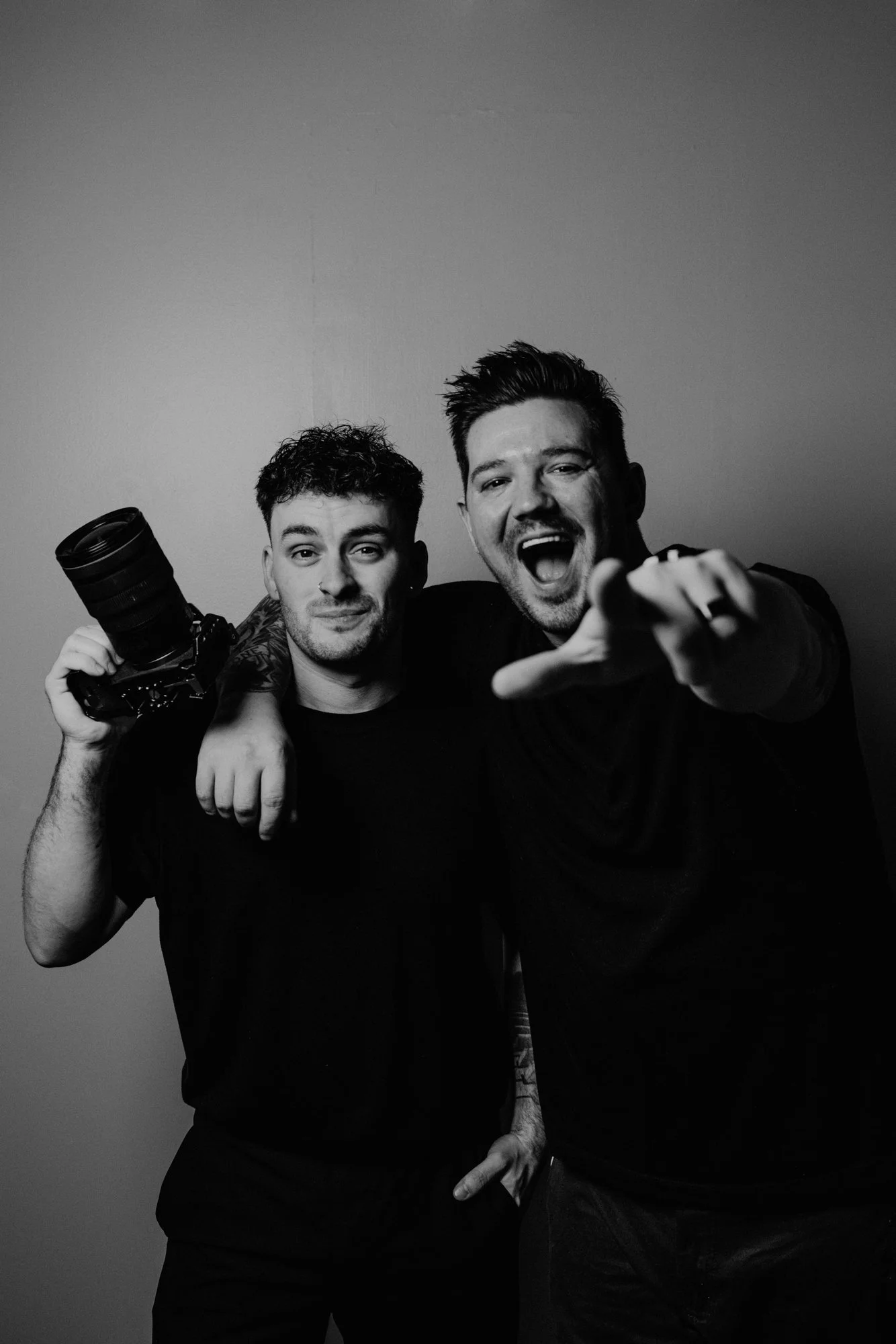 Two men smiling and posing for the camera in front of a plain background, one holding a camera and the other pointing towards the camera. The image is in black and white.