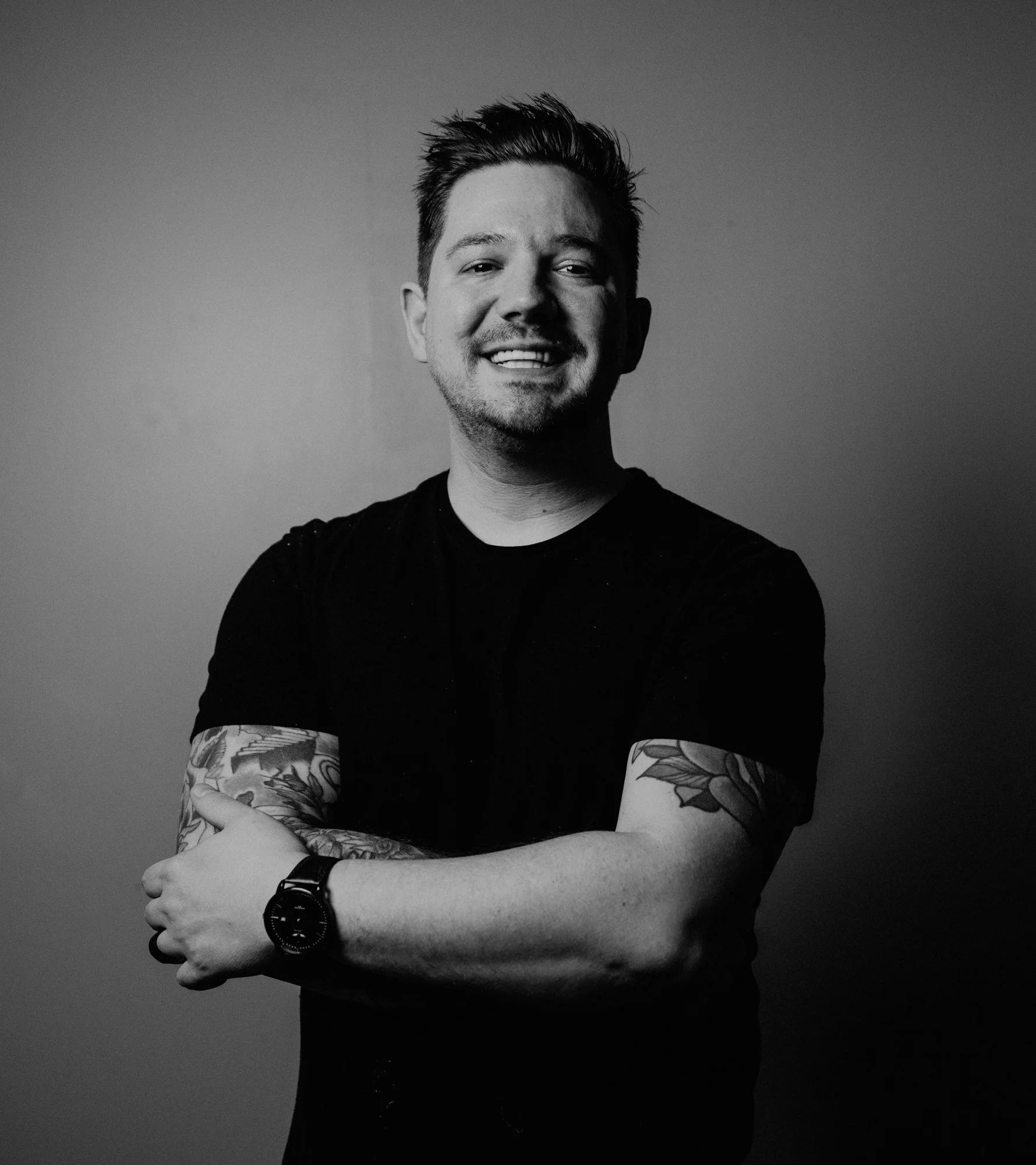 Black and white photo of smiling young man with tattoos on arms, wearing a black t-shirt and watch, crossing arms, against plain background.