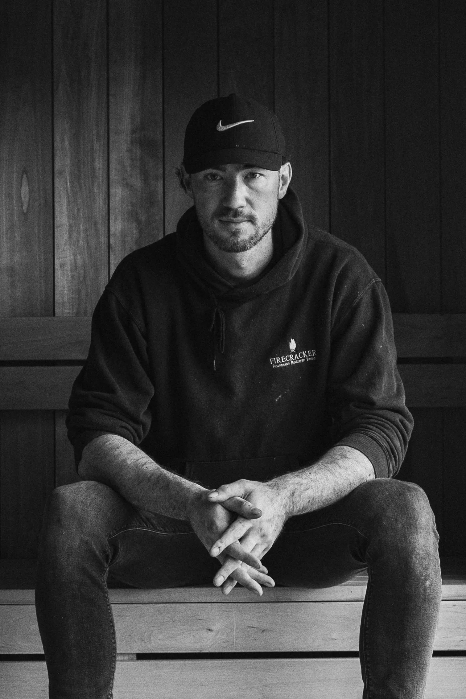 A man wearing a black Nike cap and a black hoodie, sitting on a wooden bench with his hands clasped, in front of a wood-paneled wall.
