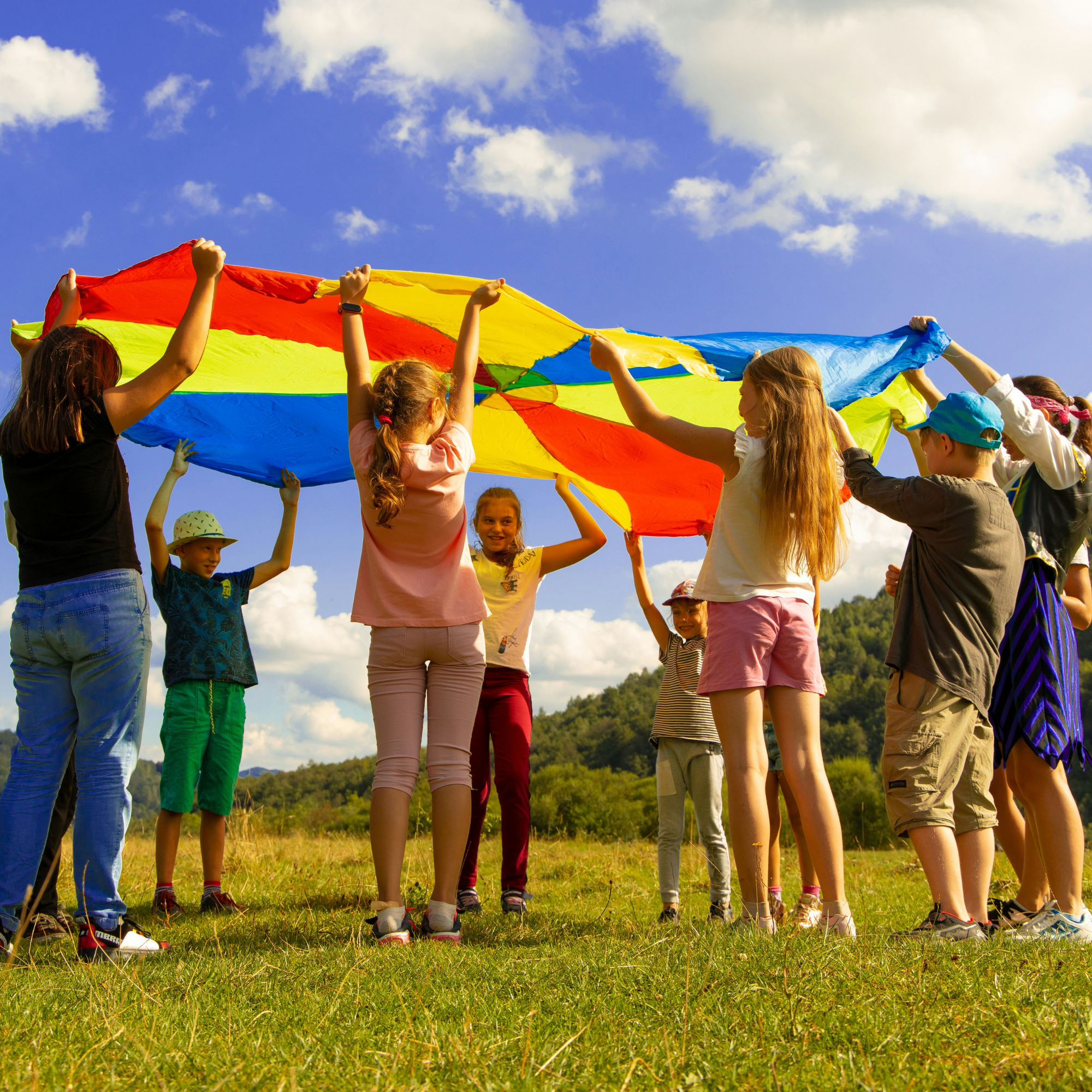 Children playing with a colorful parachute outdoors under a blue sky with clouds