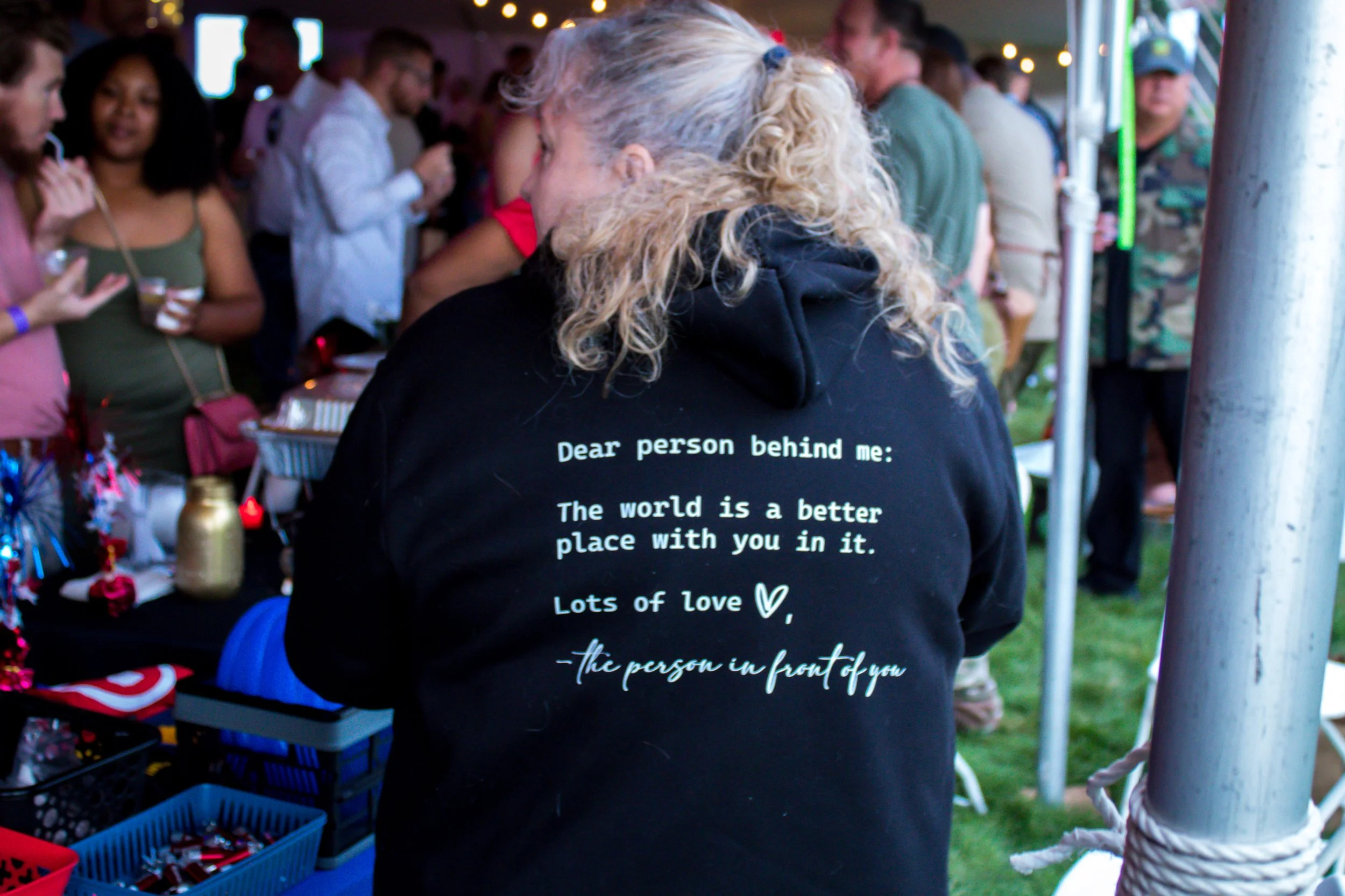 Back of a woman's black hoodie with a message that reads: 'Dear person behind me: The world is a better place with you in it. Lots of love, the person in front of you.' She is at an outdoor event with people around her and tables with items on them.
