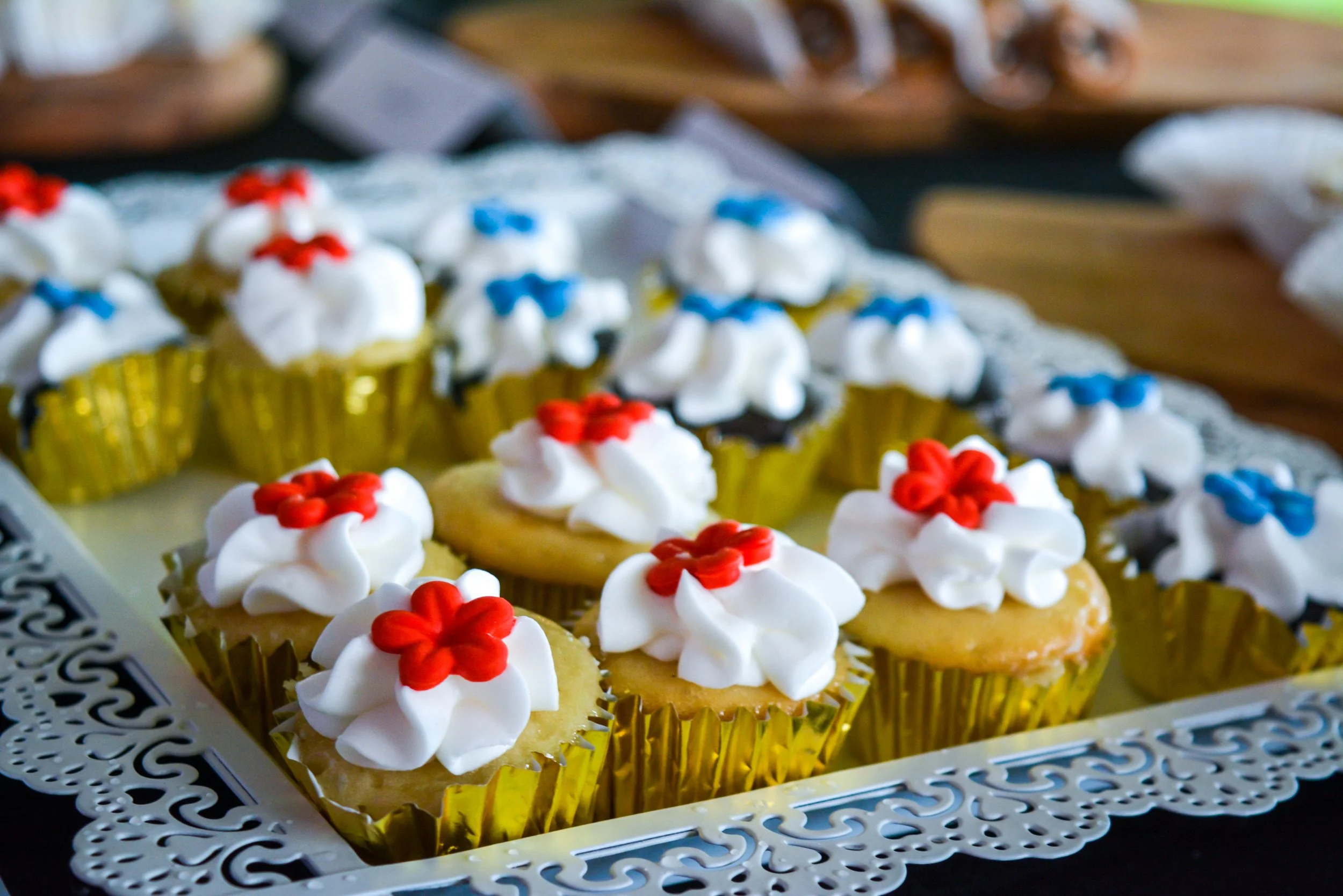 Assorted cupcakes decorated with white frosting and red or blue flower-shaped toppers, displayed on a decorative silver tray.