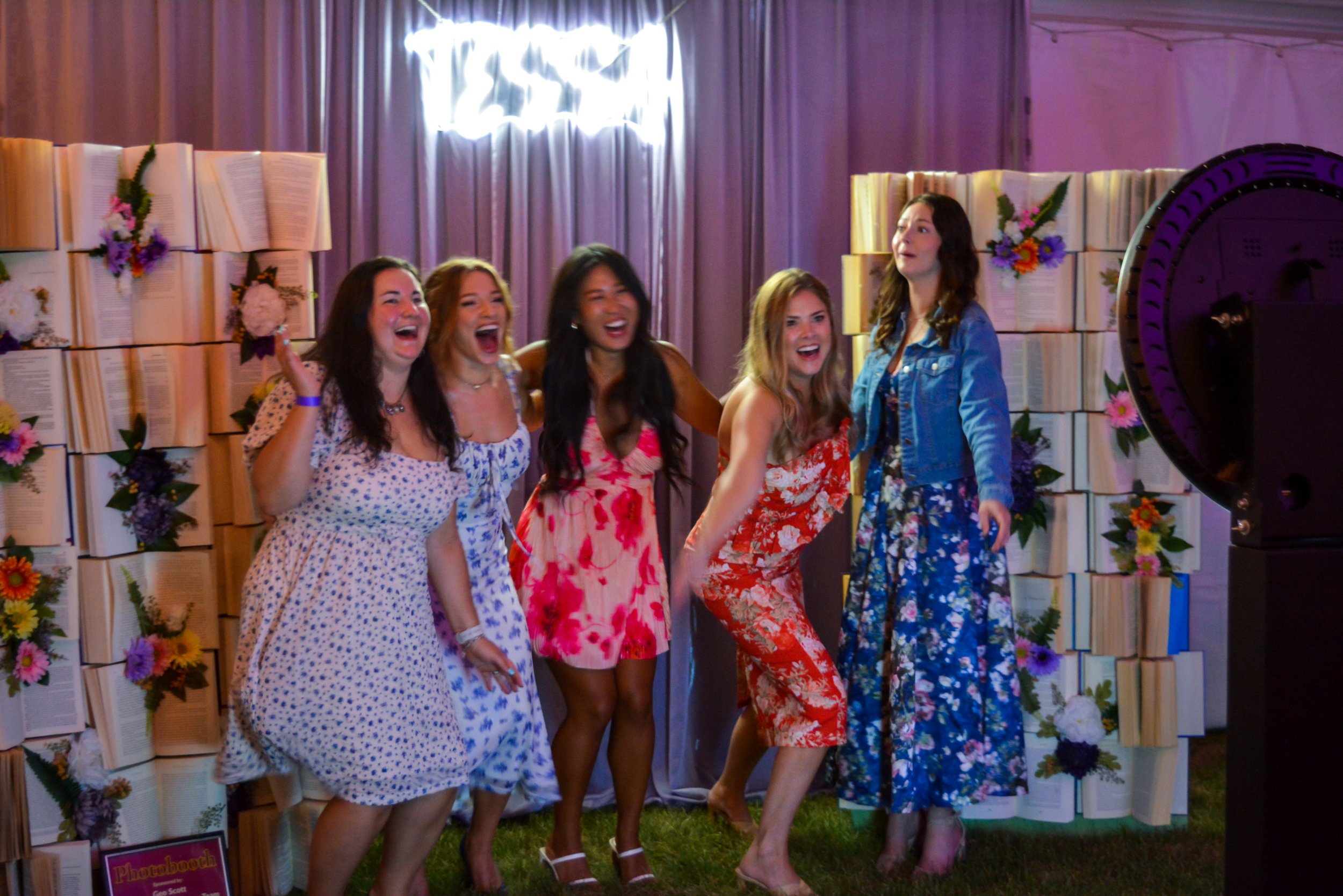 Five women in colorful dresses and a denim jacket pose and laugh in front of a decorated backdrop with books and flowers at an event.