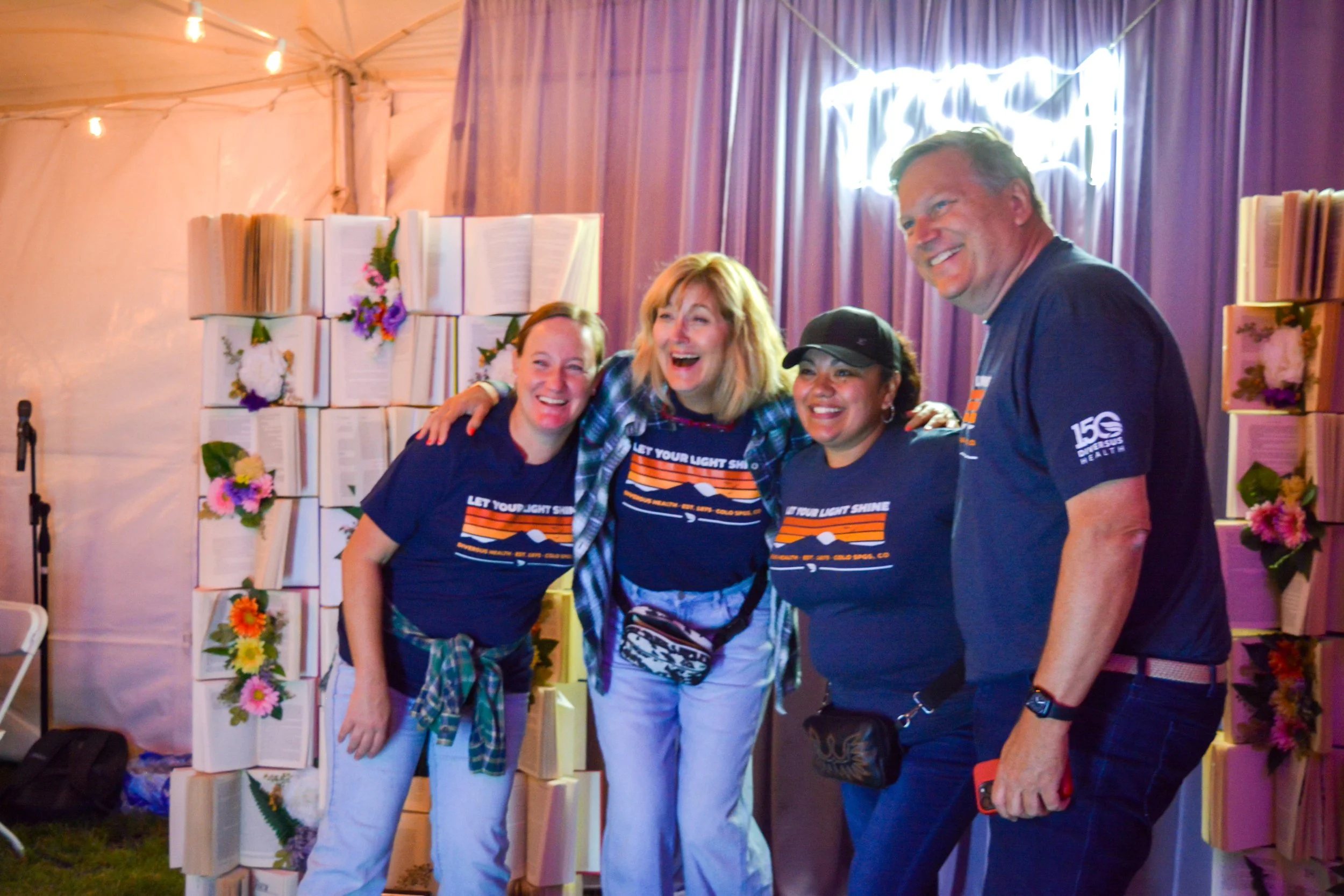 Four happy people, three women and one man, stand together in front of a decorative backdrop of books and flowers at an indoor event, smiling and posing for the camera.