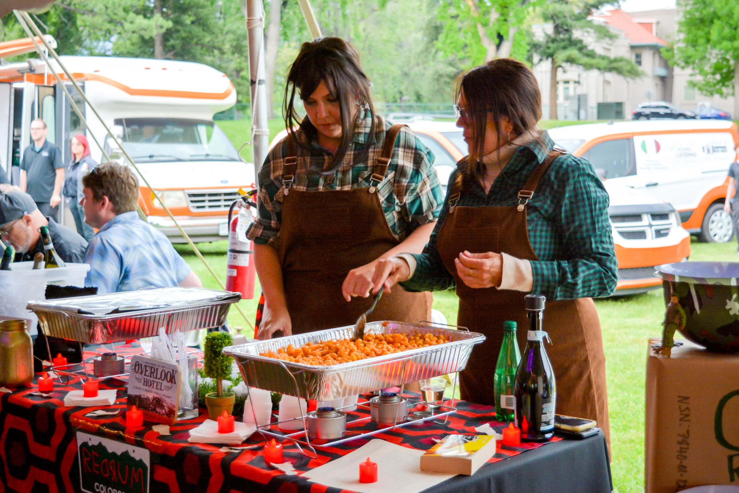 Two women wearing brown aprons serving food at an outdoor event. They are in front of a table with trays of cooked pasta, candles, and condiments. In the background, there are people sitting and standing under a tent, with vans and trees visible.