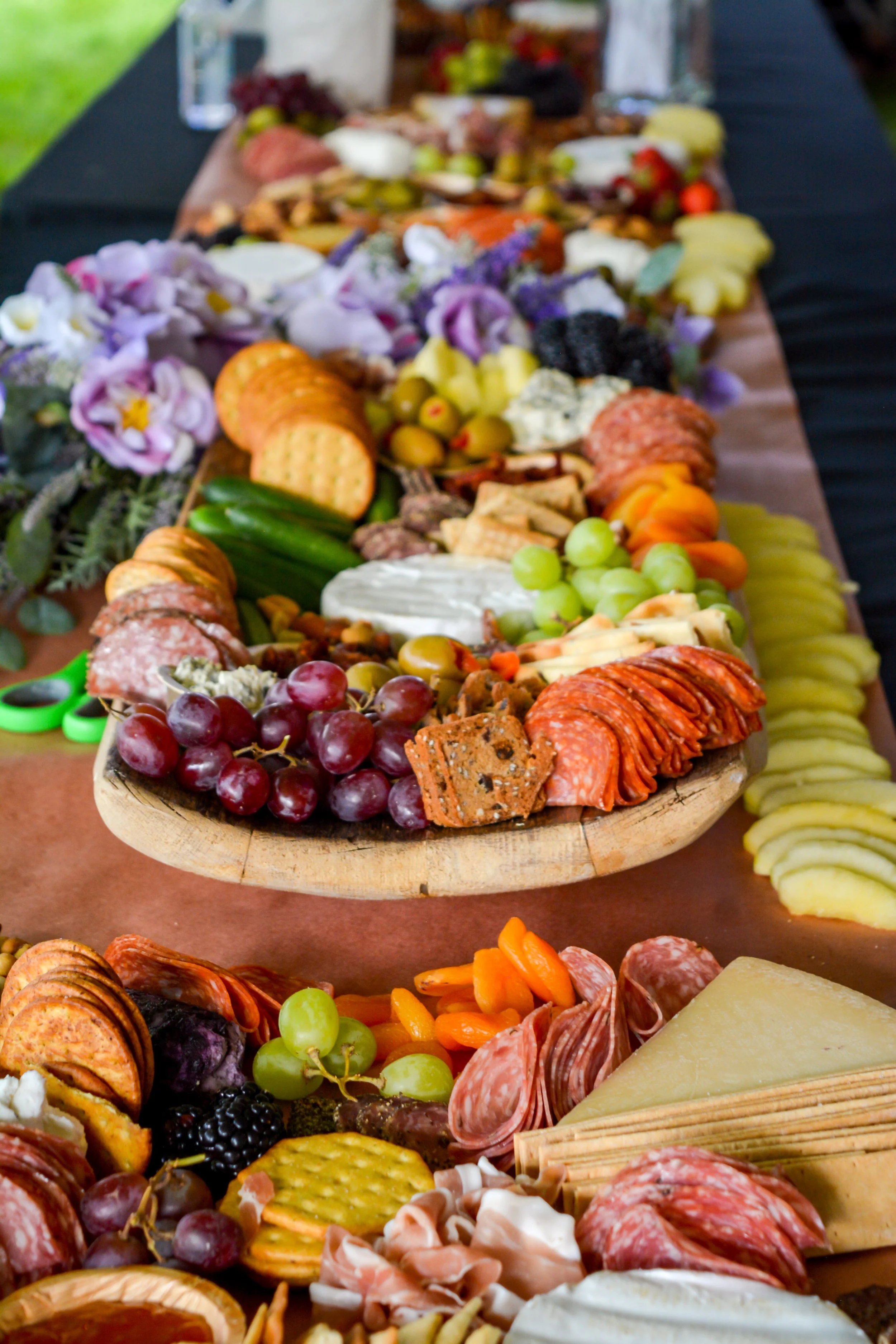 A charcuterie board featuring an assortment of grapes, crackers, meat slices, cheeses, and fruits arranged on a wooden platter with flowers.