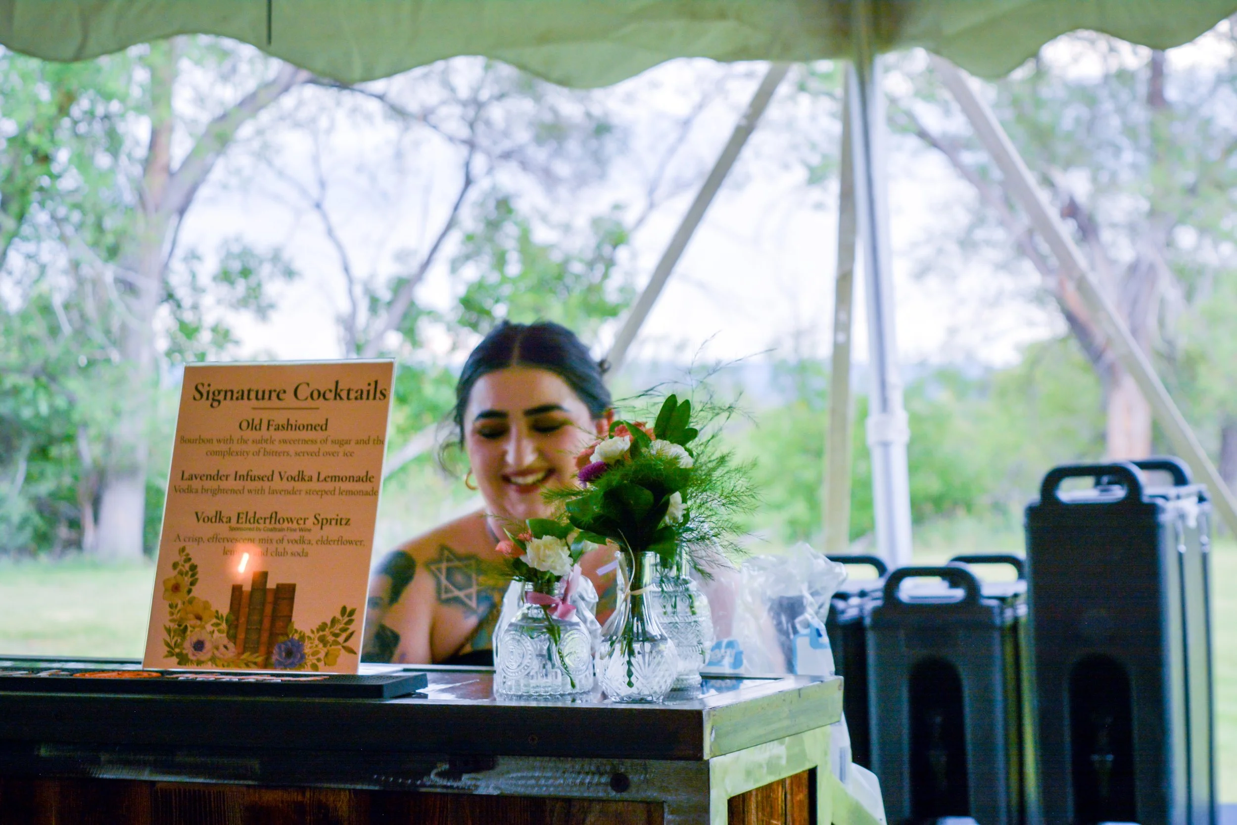 A smiling woman with dark hair and tattoos standing behind a bar, decorated with flowers in glass jars, with a sign listing signature cocktails, in an outdoor setting with trees in the background and luggage nearby.