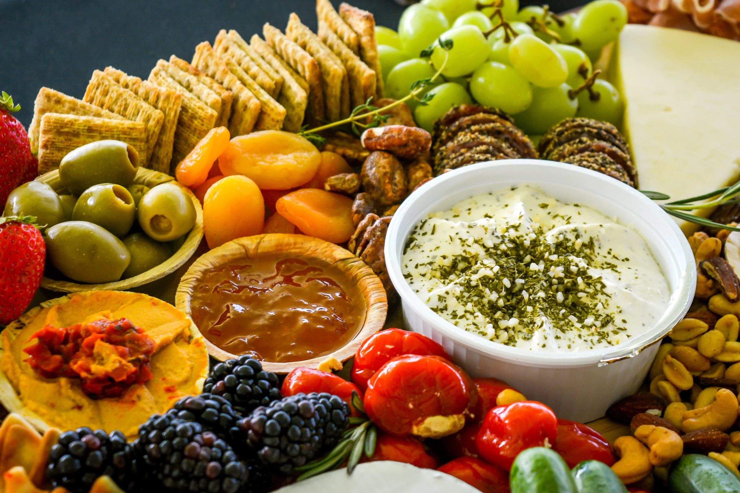 A platter of assorted snacks including green olives, strawberries, grape clusters, dried apricots, crackers, cheese, a bowl of dip garnished with herbs, and various nuts and fruits.