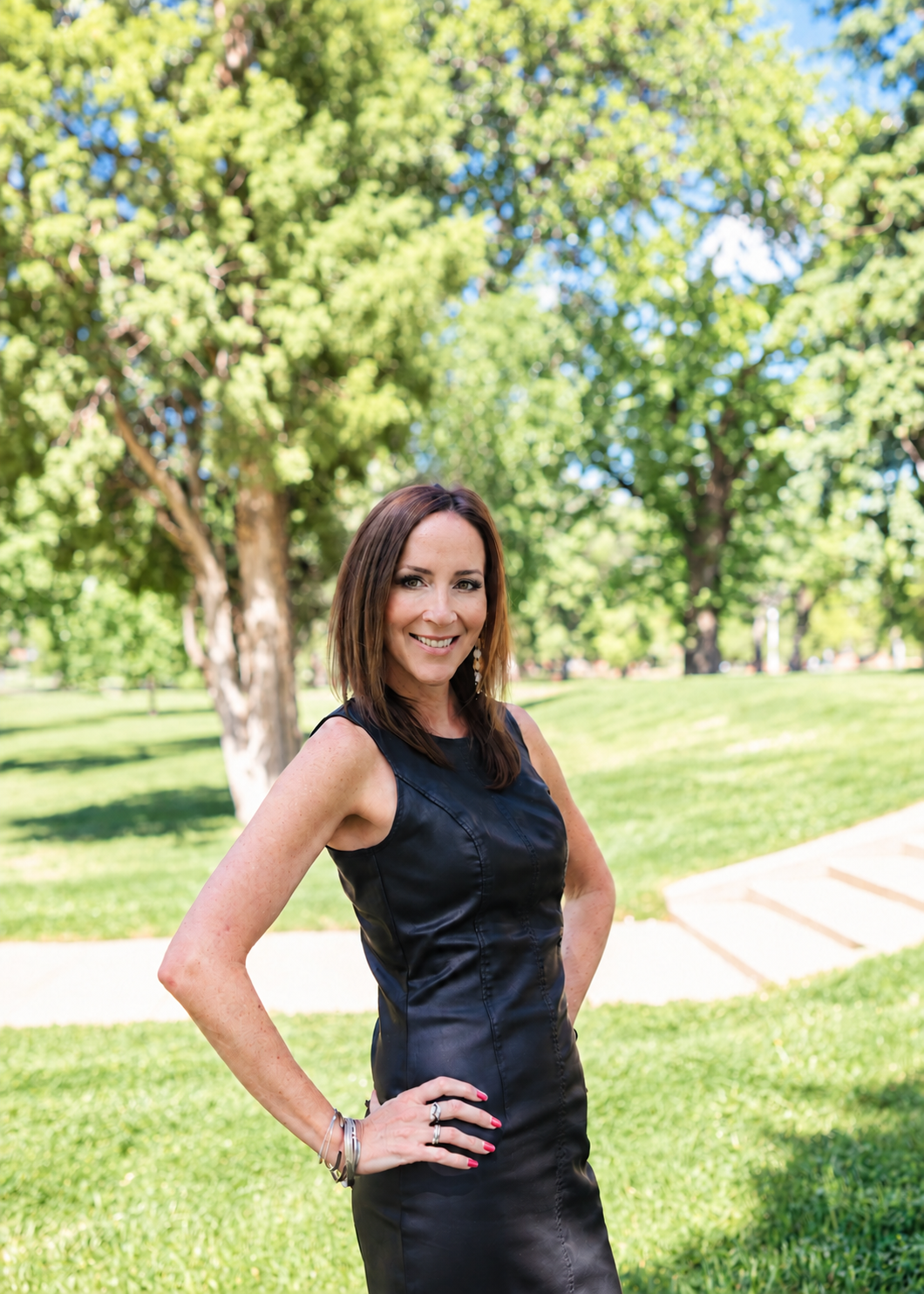 A woman with shoulder-length brown hair, wearing a black blazer and statement earrings, standing outdoors with a green, leafy background. She is smiling and has her left hand on her hip.