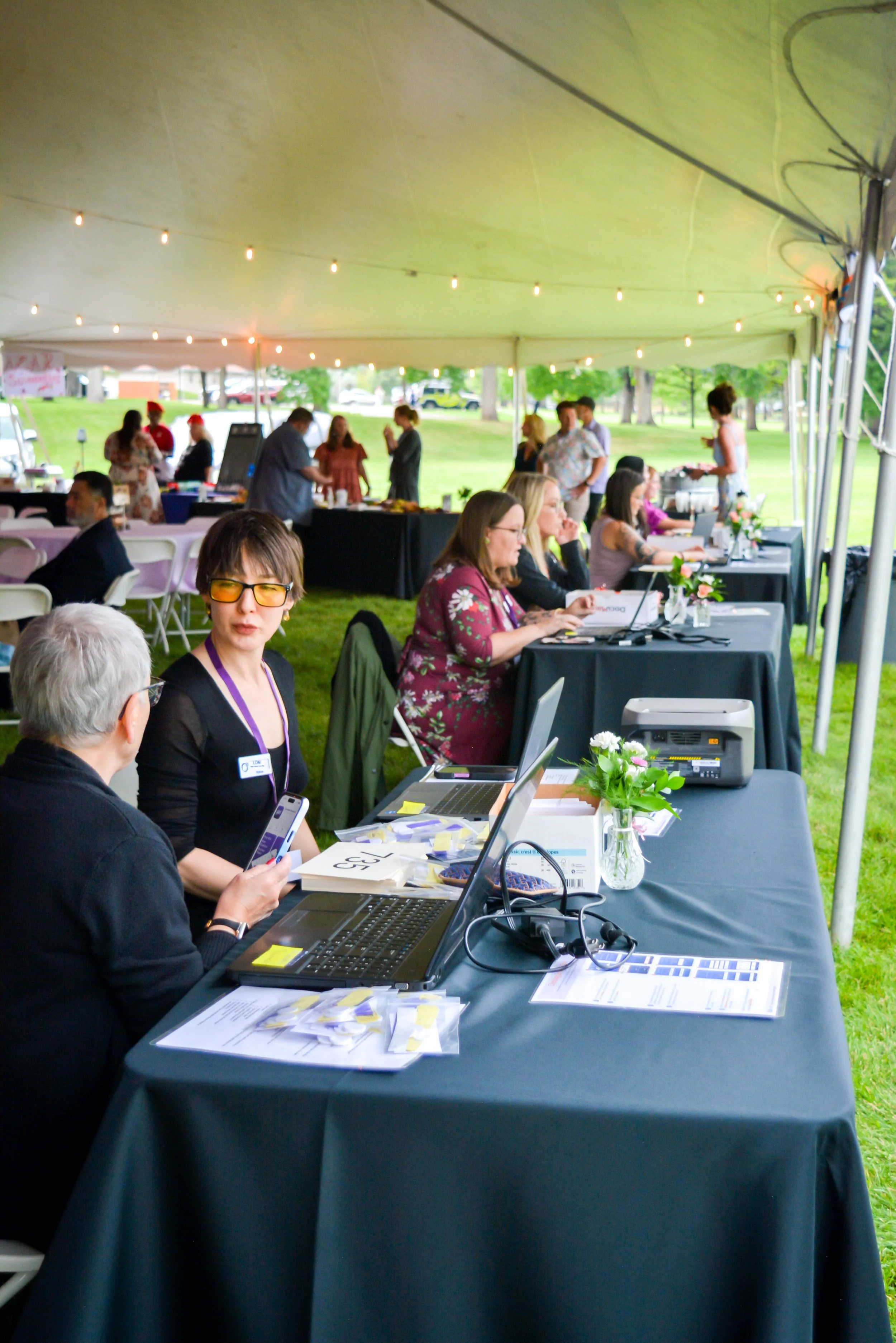 People sitting at a registration or check-in table under a large white tent with string lights and a park in the background, with some people standing and chatting in the distance.