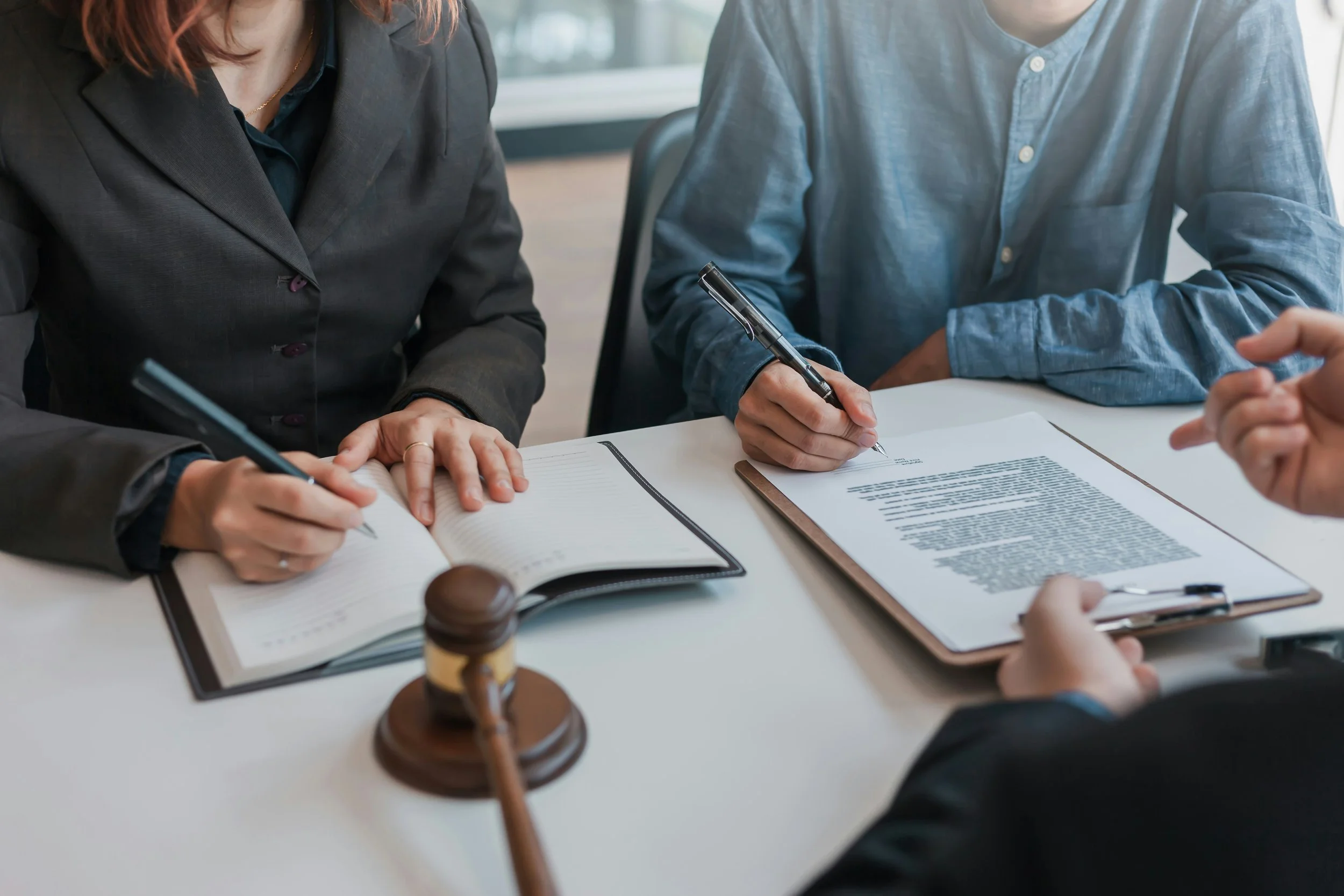 Legal professionals conducting a meeting, with one person holding a pen and capturing notes, seated at a table with legal documents, a gavel, and a hand gesturing during the discussion.