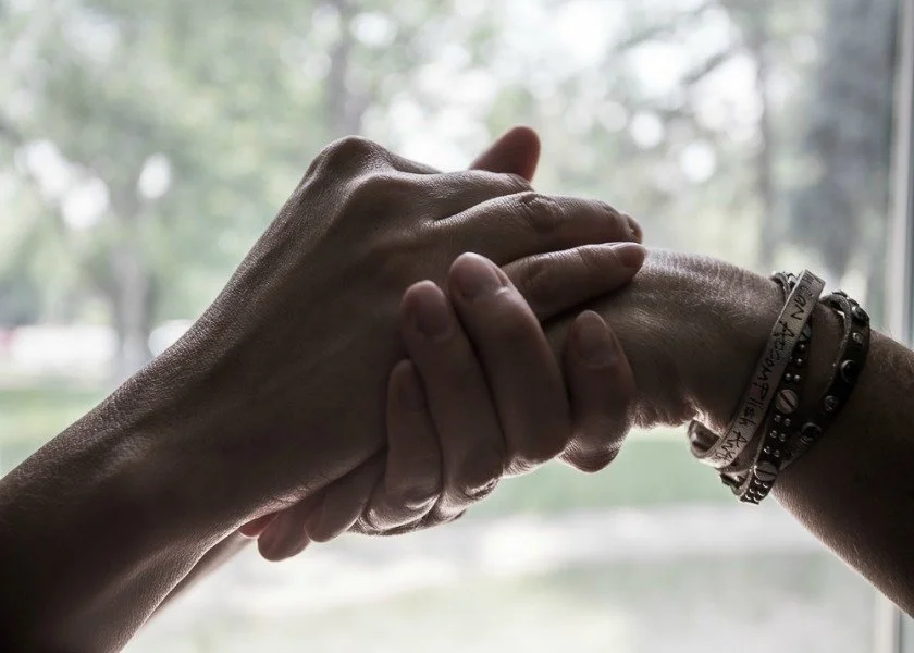 Two hands clasped together in a handshake, one wearings multiple bracelets, with a blurred outdoor background.