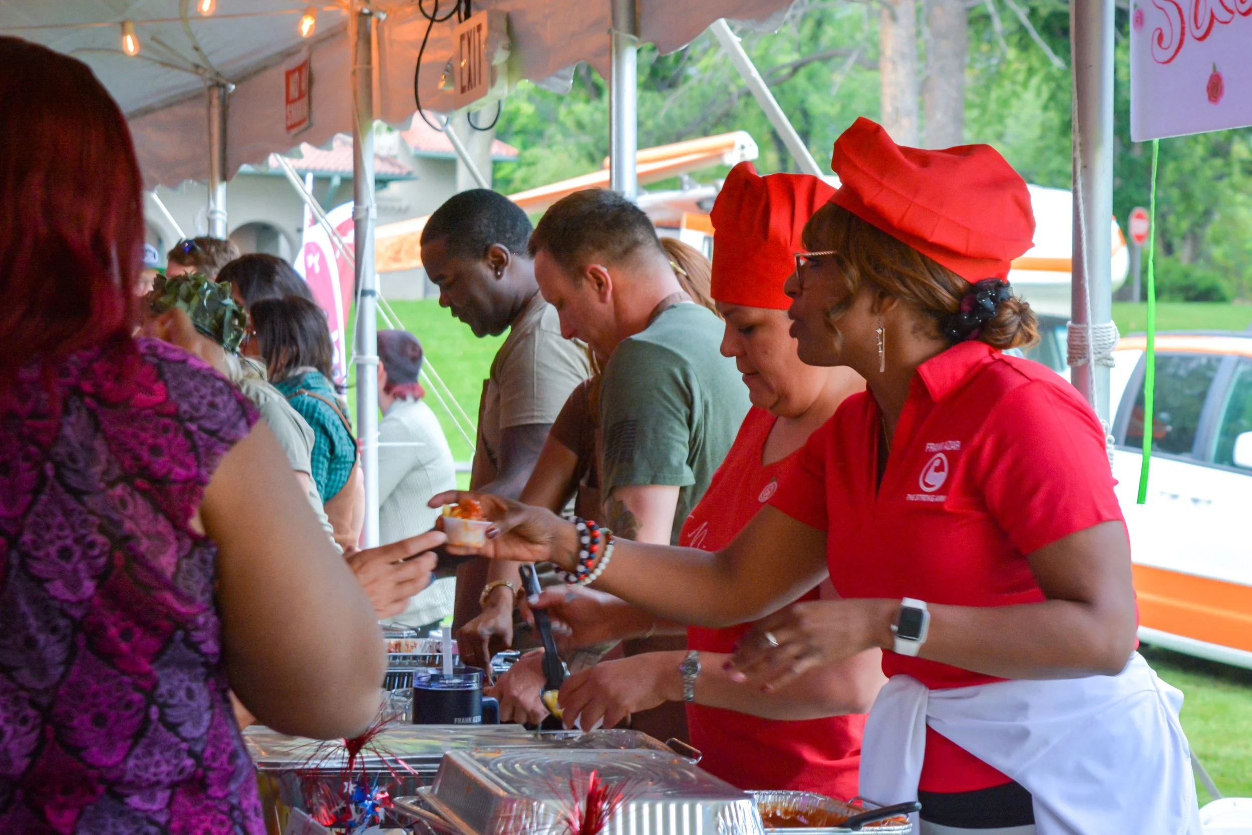 People serving food at an outdoor event booth, with some wearing red uniforms and head coverings, under a white tent. Green grass and trees are visible in the background.