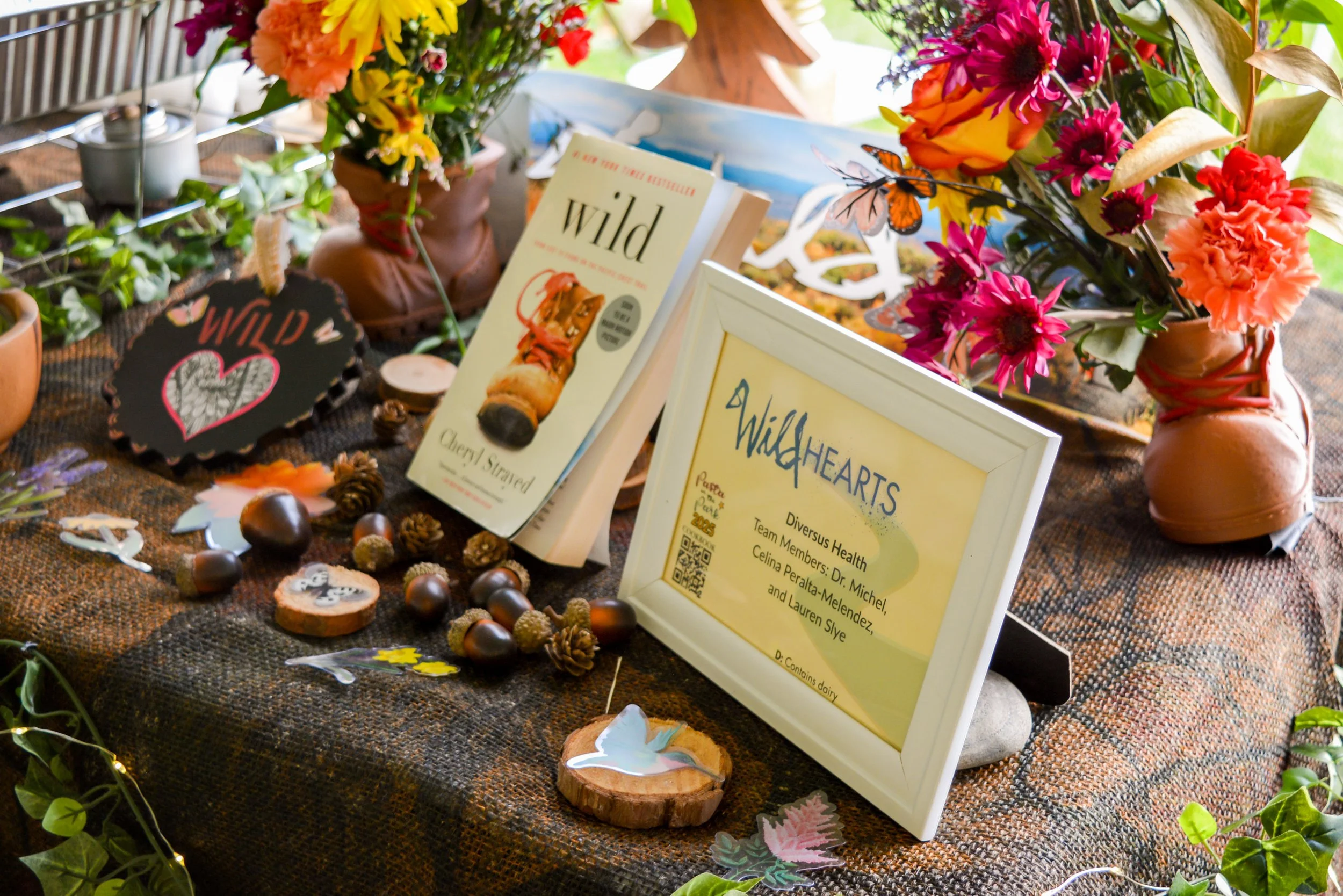 A decorated table with vases of colorful flowers, framed signs, books, acorns, pinecones, wooden decorations, and paper butterflies. The signs mention 'Wild Heart' and 'Wild' by Cherry Strayed, as well as a certificate of recognition for 'Wild Hearts' team members.