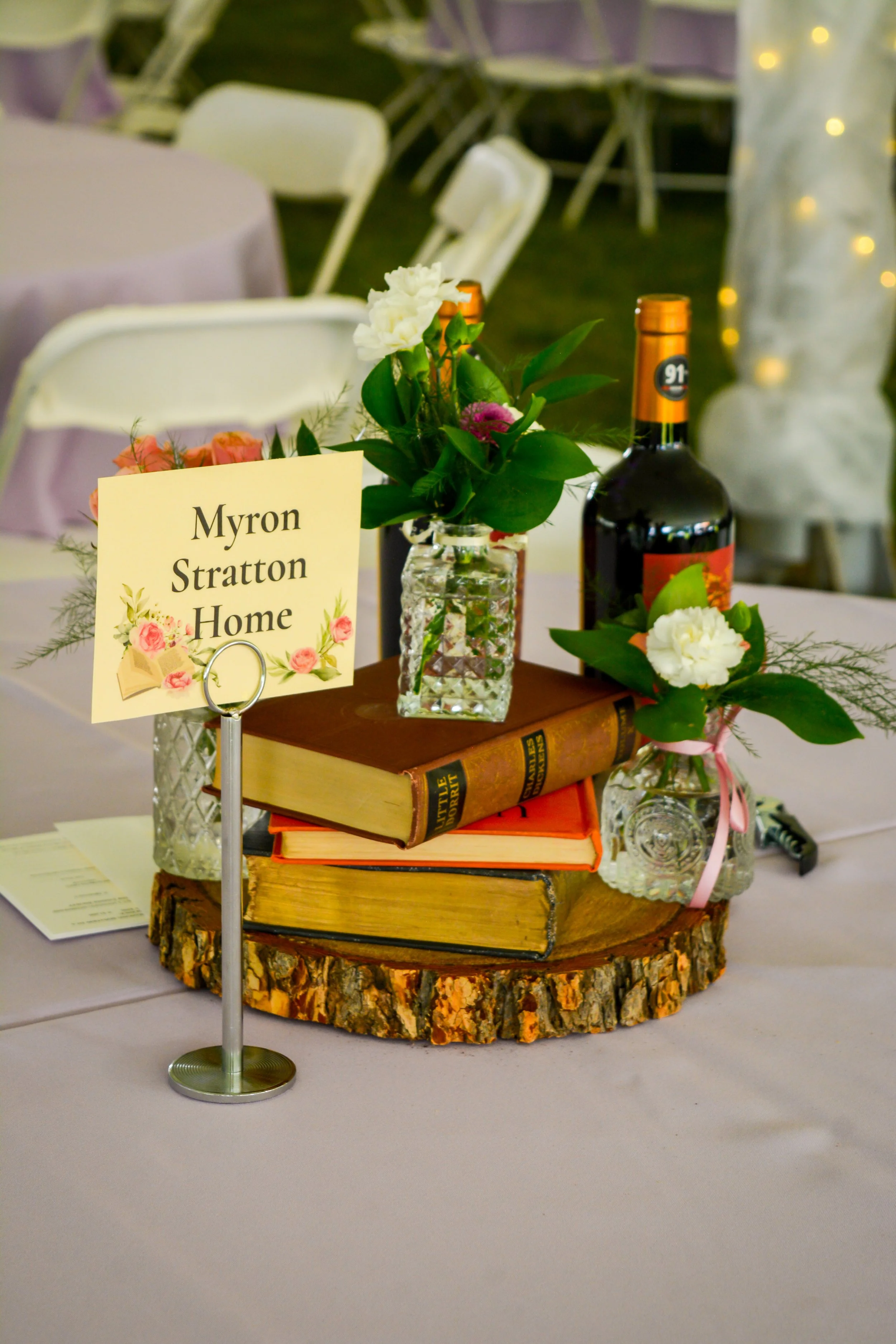 A decorated table centerpiece featuring flowers in glass vases, stacked vintage books, a bottle of wine, and a sign that reads 'Myron Stratton Home', on a wooden round base.