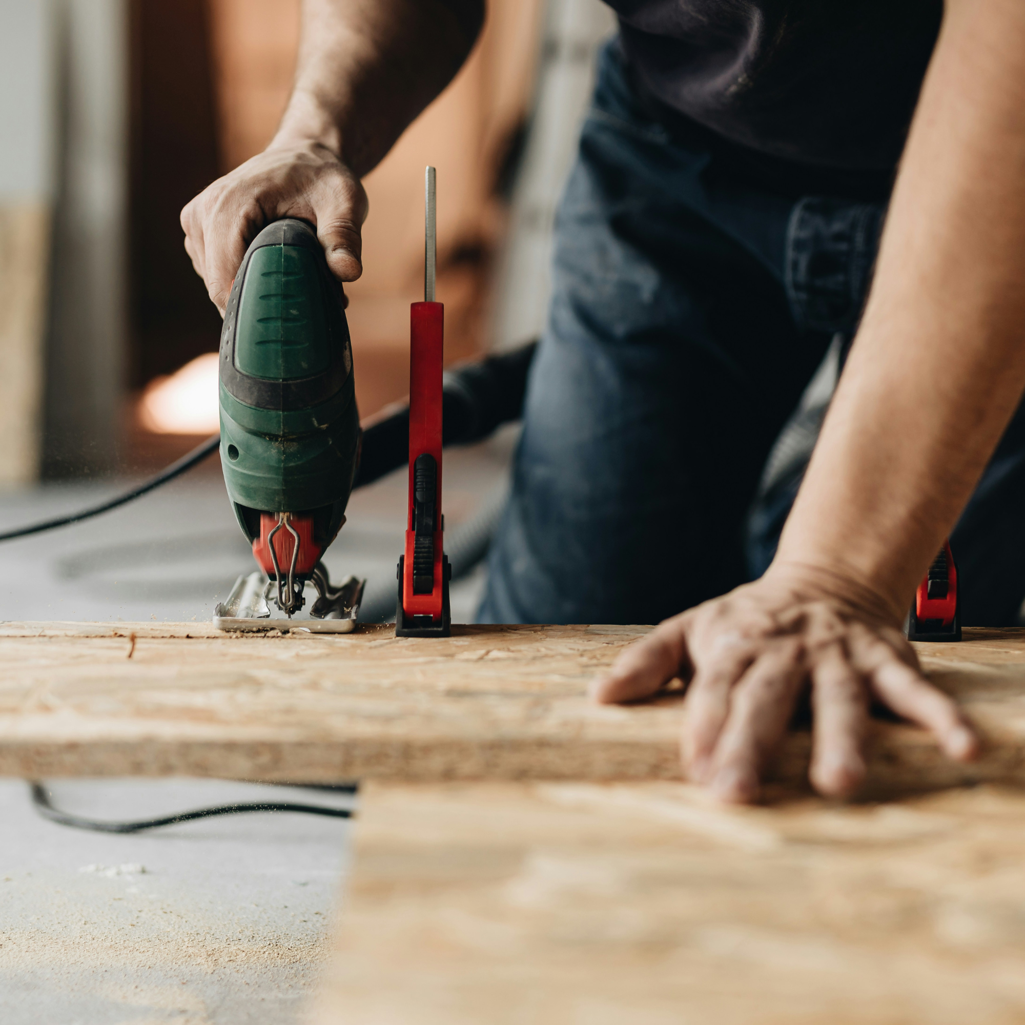 A person cuts a wooden board using a handheld electric jigsaw, with a red carpenter's square and a marker nearby.