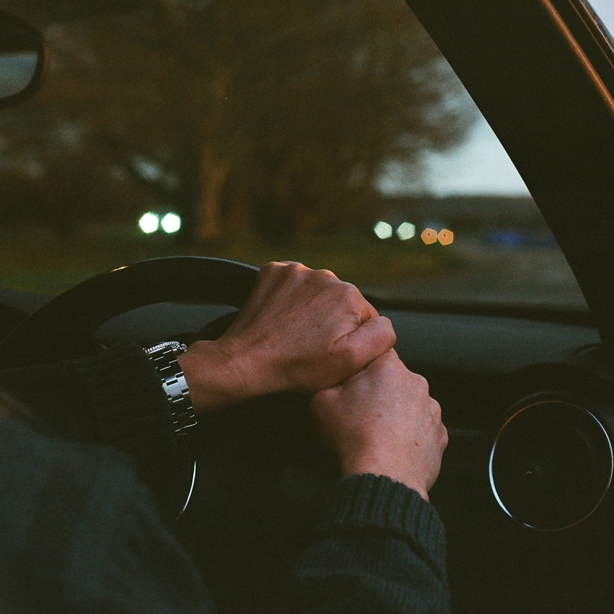 A person driving a car at dusk or night, with hands on the steering wheel, wearing a silver watch and a dark long-sleeve shirt or sweater.