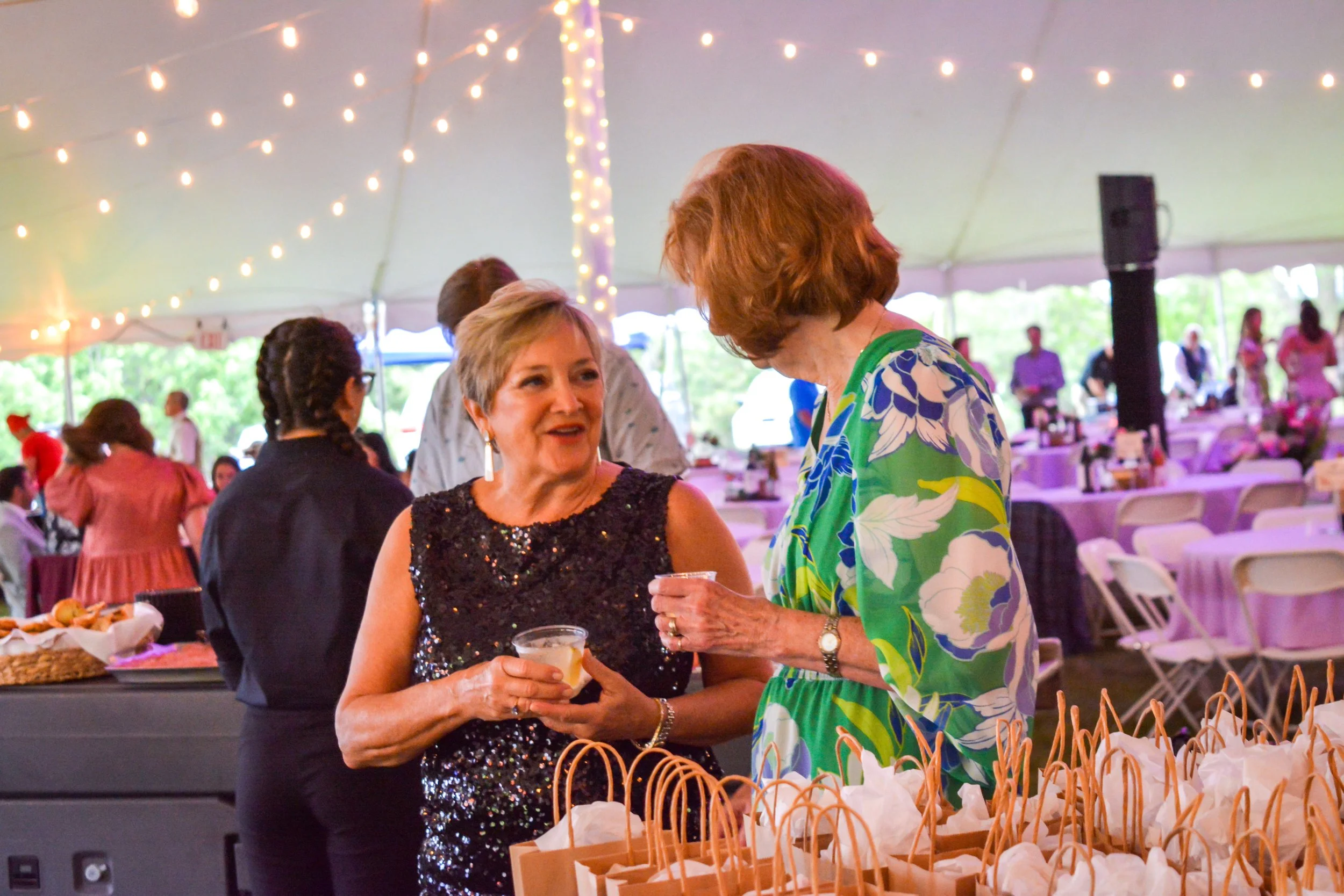 Two women conversing at a reception under a large tent with string lights. One woman in a sparkly black dress holds a drink, while the other in a colorful floral dress holds a small cup. Gift bags are on the table in front of them.