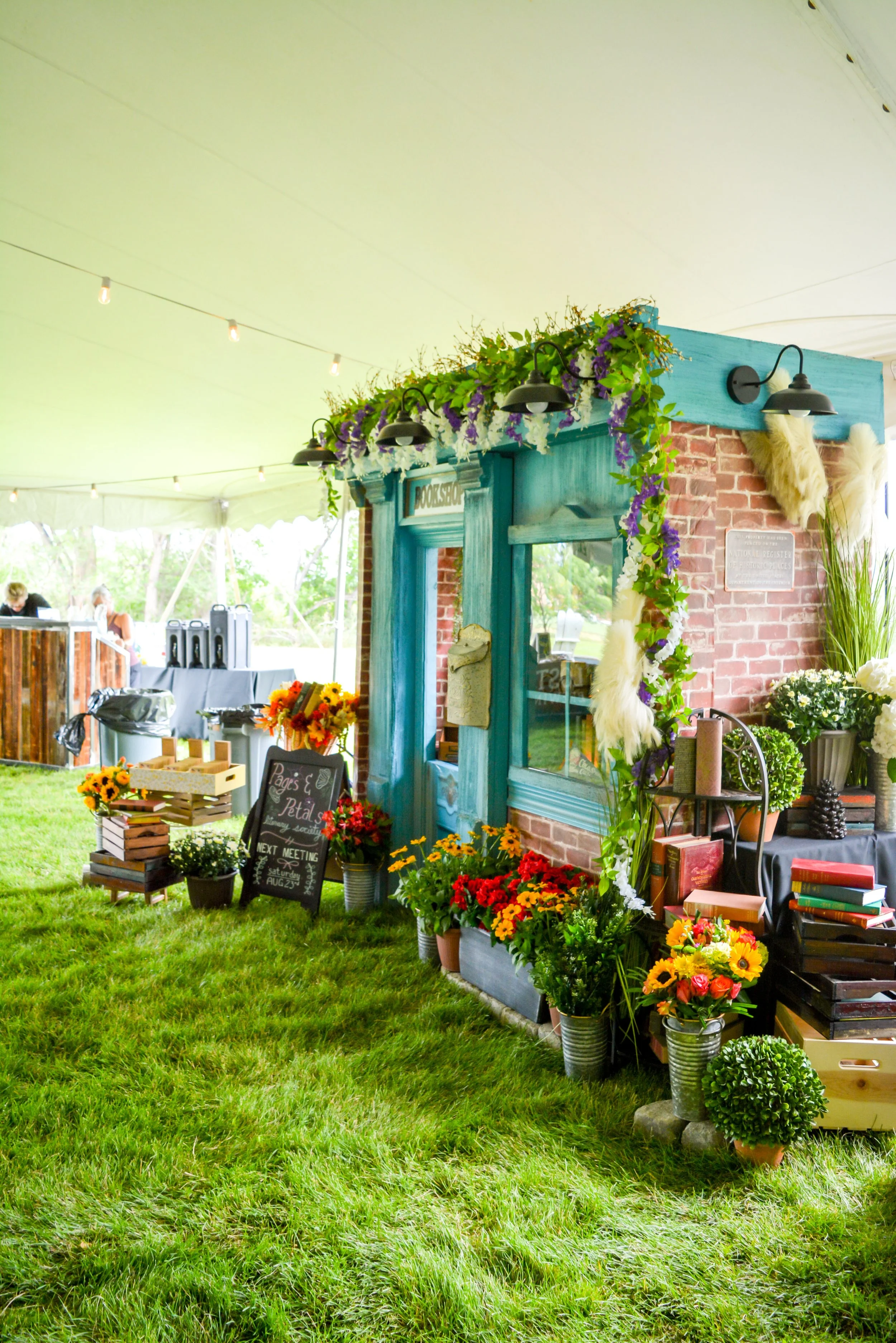 Colorful flower shop setup under a tent with various potted plants and flowers, a chalkboard sign, and decorative lighting.