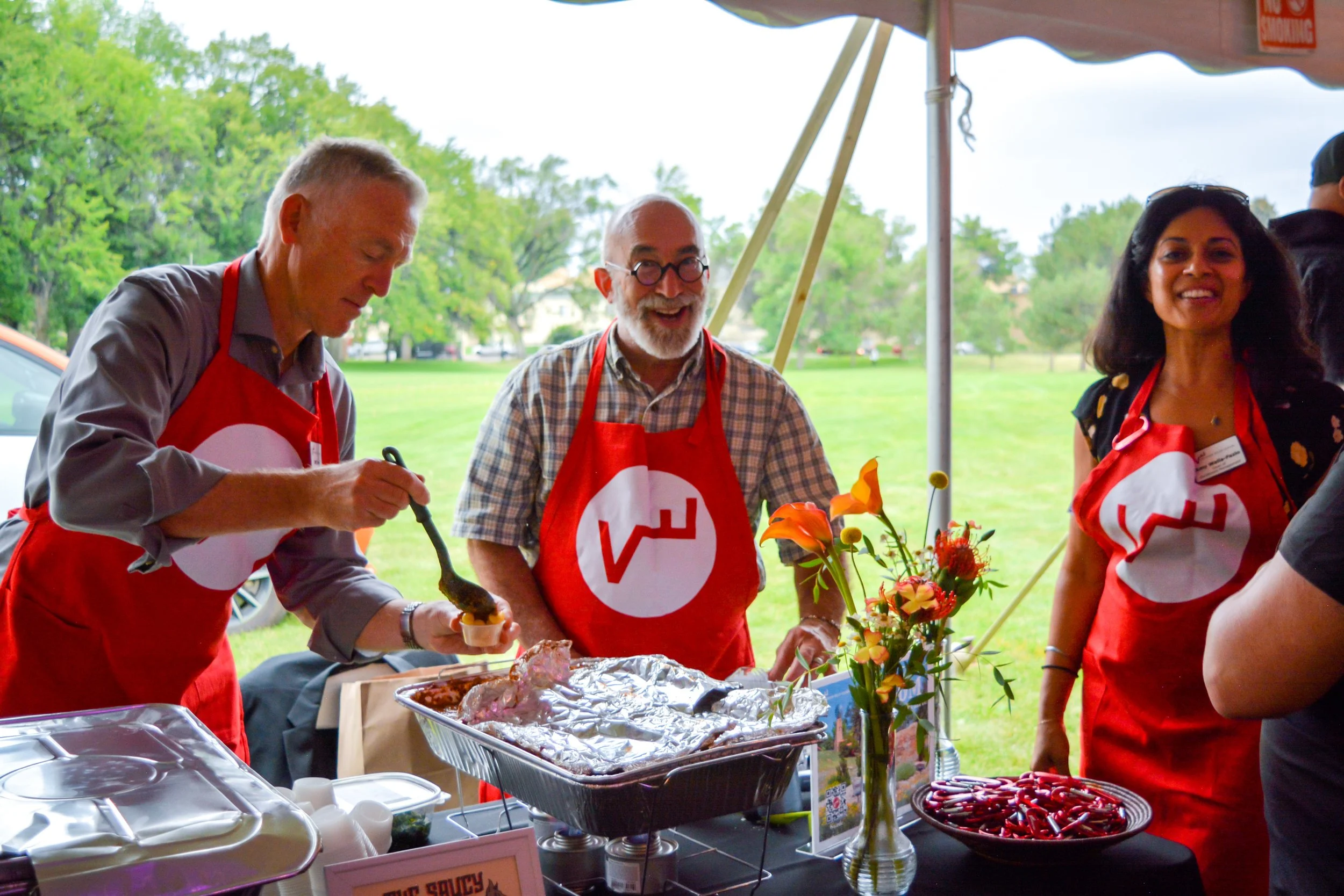 Three people wearing red aprons with a white logo, working at a table under a tent outdoors with green trees in the background. One man is serving food, the woman is smiling, and another man is looking down at his food. The table has various food items, a flower vase, and a bowl of red chili peppers.