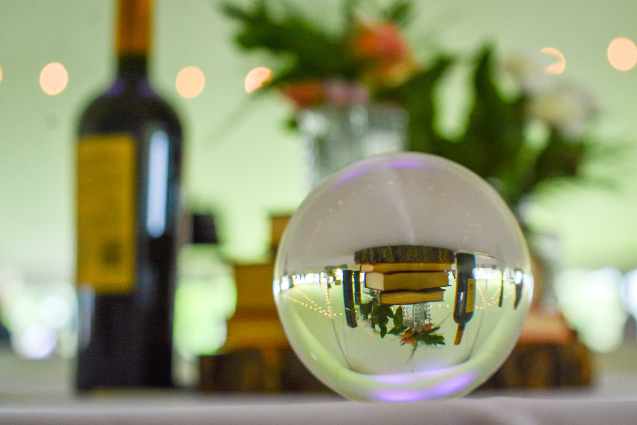 A decorative glass sphere reflecting a blurred background of a table with books, a bottle, and a vase with flowers in a room with green walls and string lights.