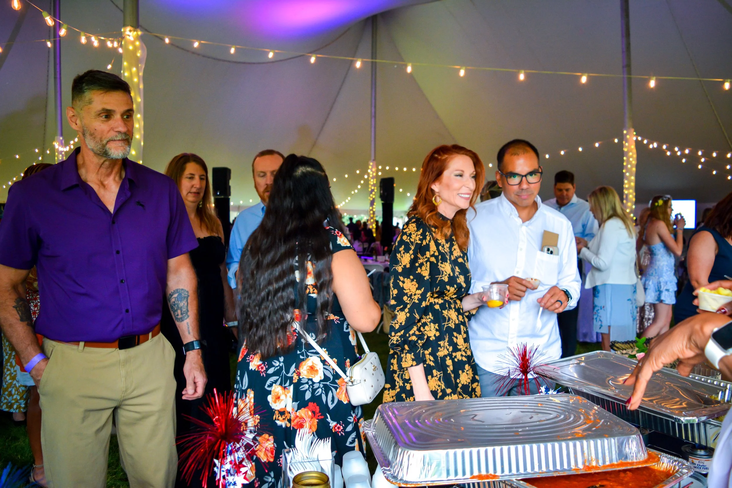 People gathered at a buffet table during a celebration in a decorated tent with string lights.