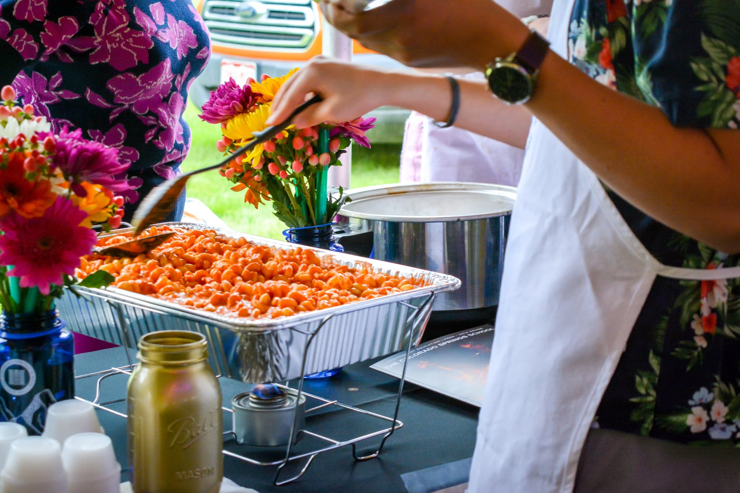 Someone serves food at an outdoor event, with a tray of macaroni and cheese, colorful flowers in a vase, and a person wearing a white apron.