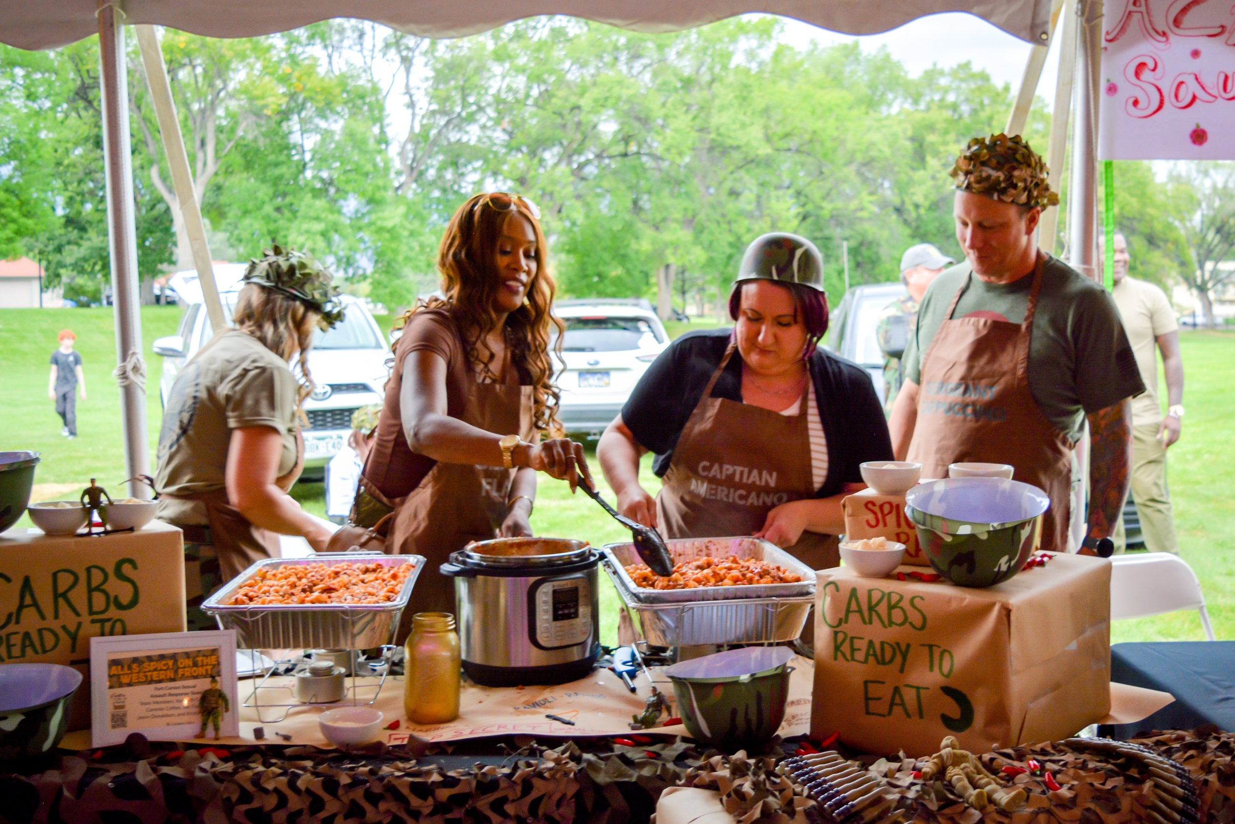 People dressed in camouflage clothing and wearing aprons with military titles, serving food at an outdoor event, with trees and cars in the background.