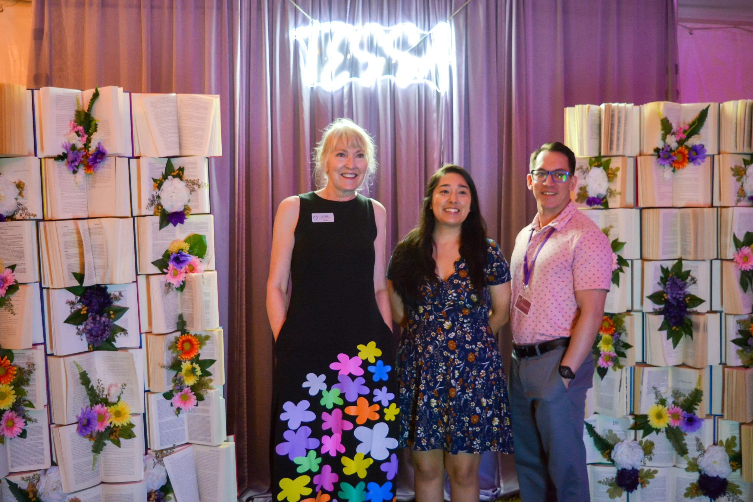 Three people standing in front of a decorative backdrop made of books and flowers, smiling for a photo at an indoor event.