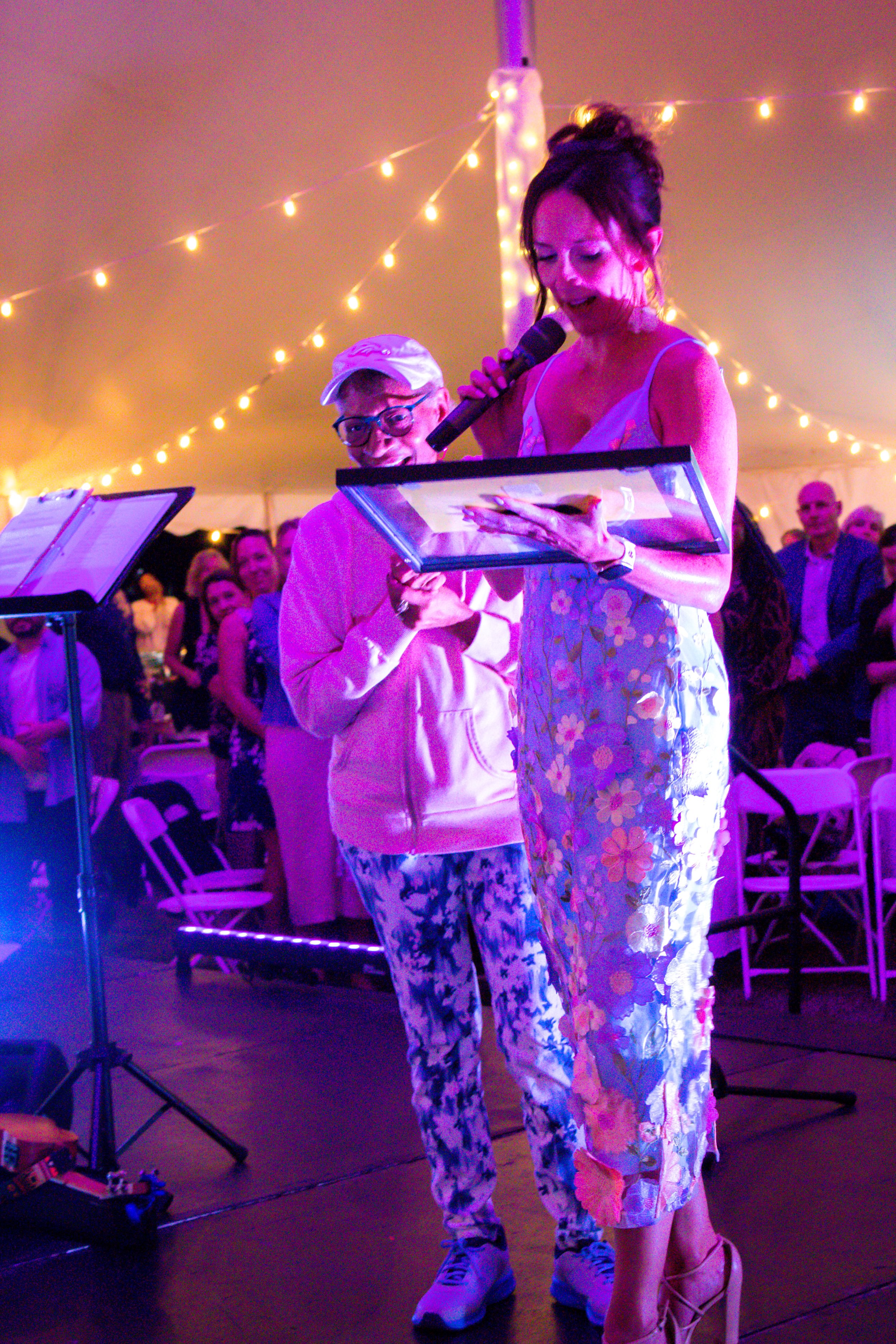 A woman in a floral dress reads from a framed item while speaking into a microphone at a lively event under warm string lights, with a smiling person in casual attire standing nearby and an audience in the background.