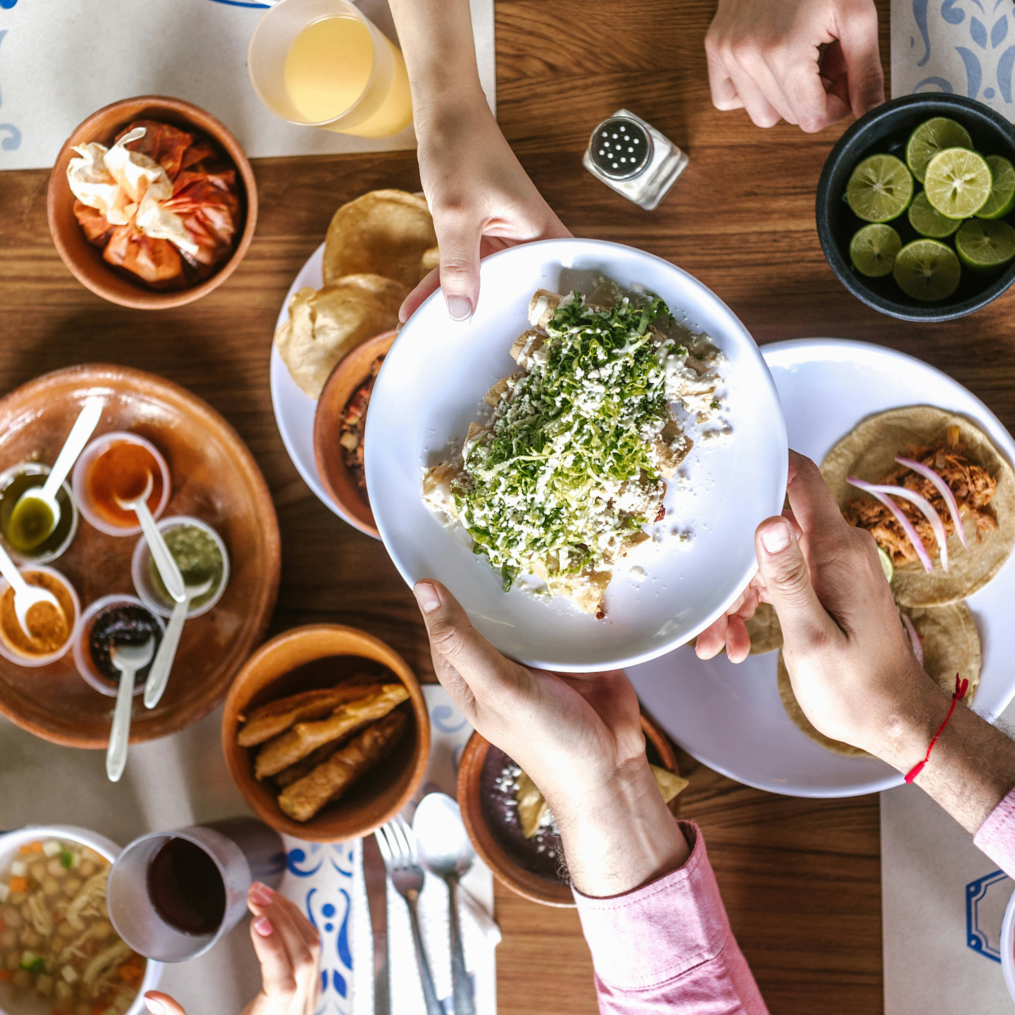 People sharing Mexican food including tacos, guacamole, and salsa, with lime slices and drinks on a wooden table.