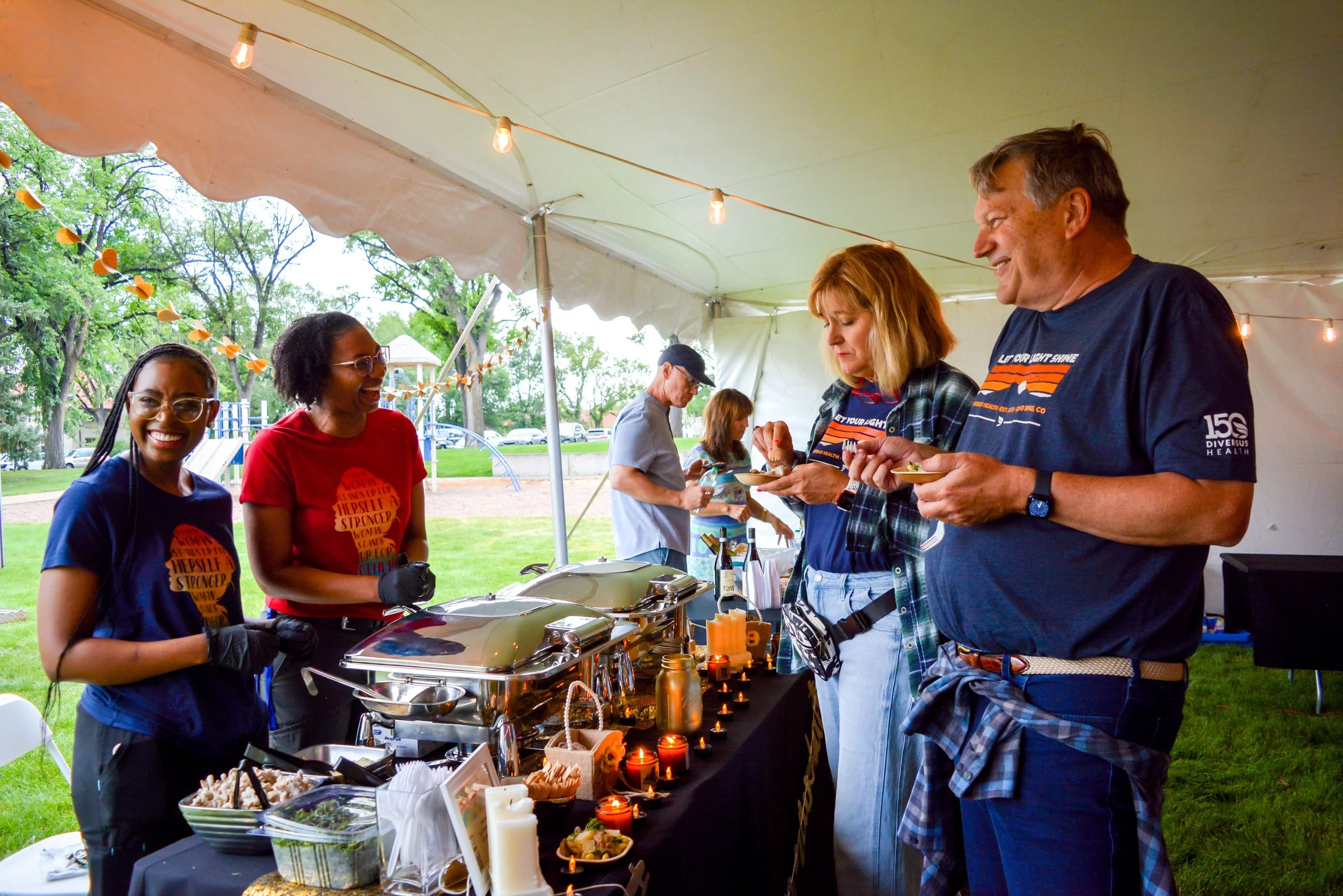 Group of people enjoying a food festival under a tent, with two servers in front and people choosing food from the buffet table.
