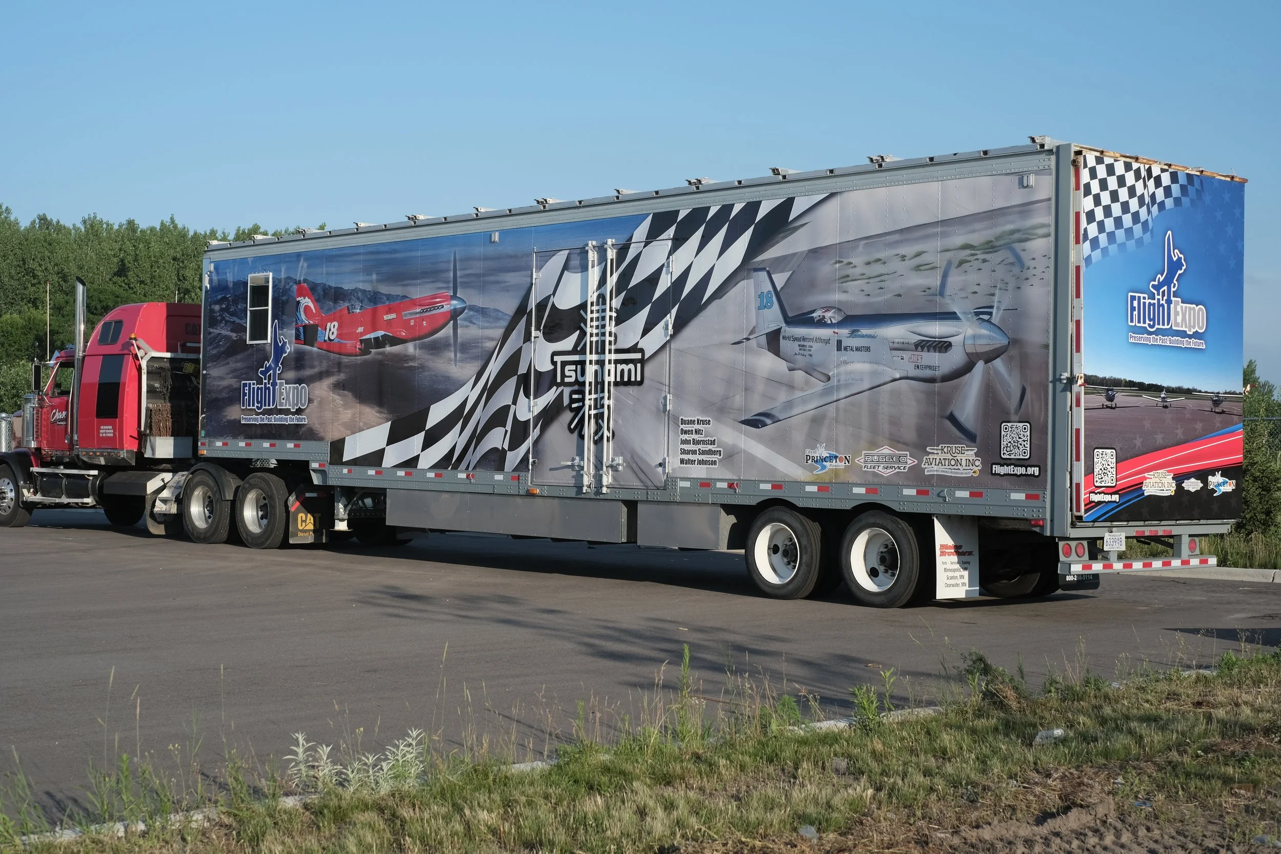 A semi-truck with a colorful design featuring an F-16 fighter jet and the logo for Flight Expo, parked outdoors with trees and a clear blue sky in the background.