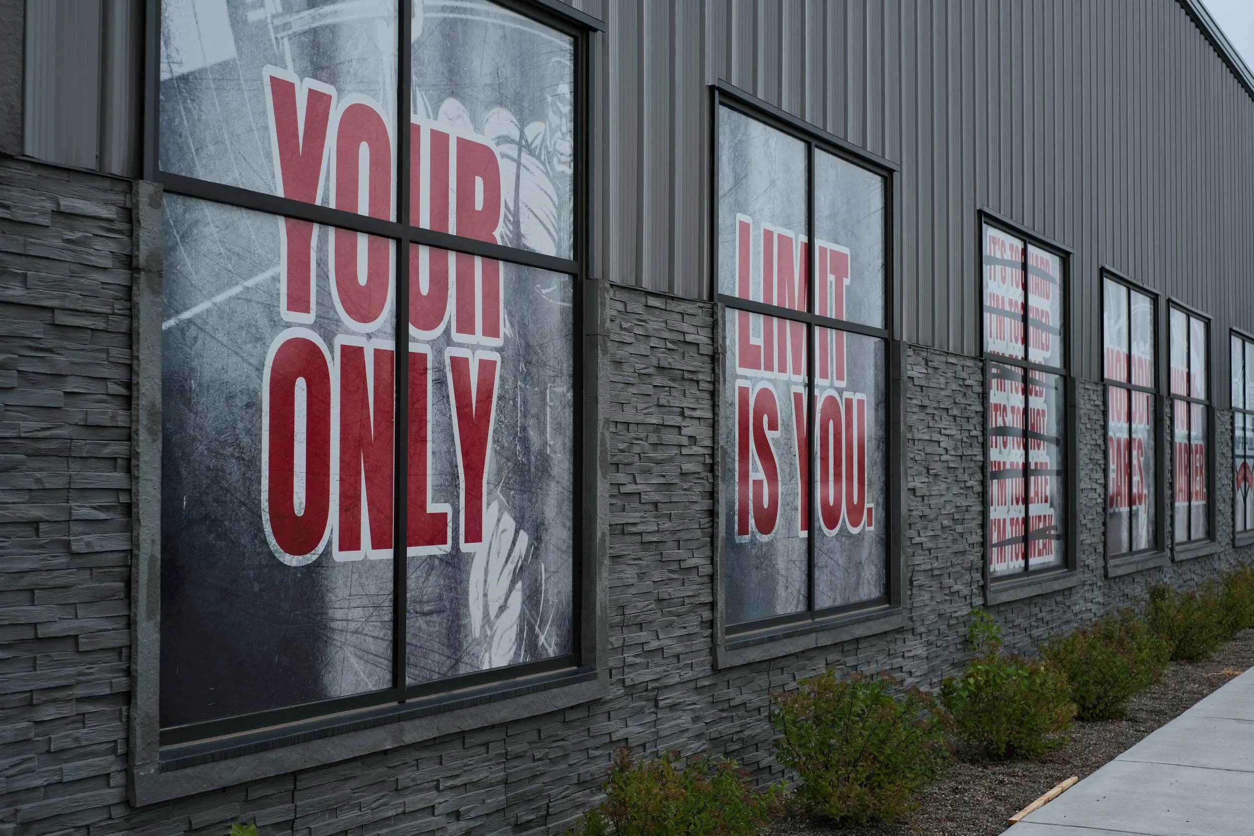 The exterior of a building with large windows displaying signs that say 'YOUR' and 'LOVE' in red letters on white backgrounds. The building has gray siding and a stone accent wall below the windows. There are small plants and a sidewalk along the bui