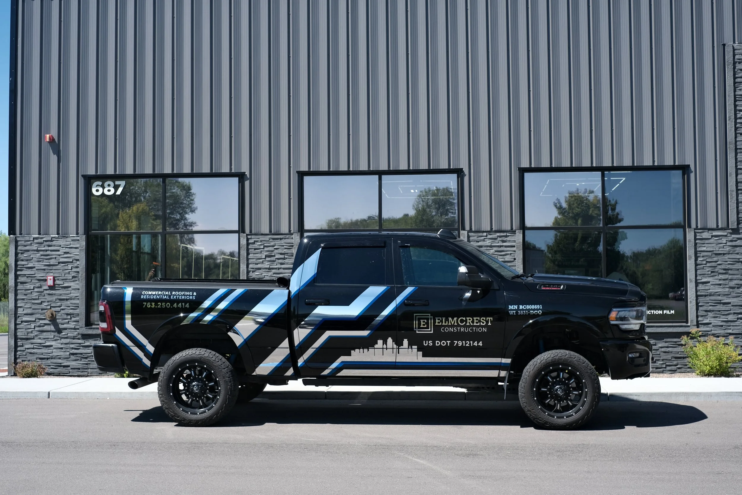 A black pickup truck with blue and white stripes parked in front of a commercial building with large windows and gray metal siding. The truck has company information printed on the side, including a phone number and construction services. The buildin