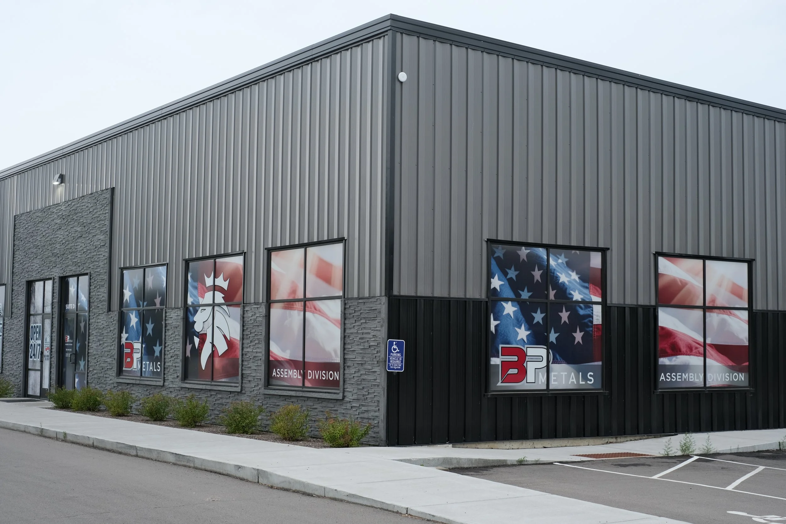 Exterior view of a commercial building with large windows displaying patriotic American flags and banners for B&P Metals with an assembly division. The building has a gray metal siding with a corner accent and a small parking sign visible near the en