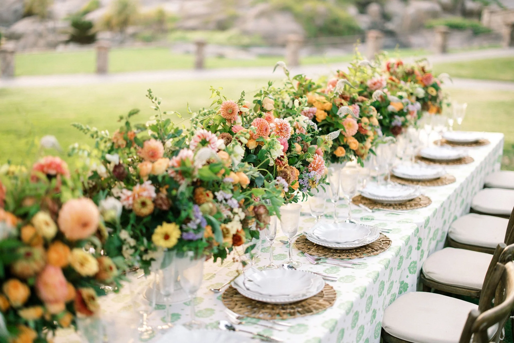 Outdoor banquet table decorated with a large colorful flower arrangement and neatly arranged plates, glasses, and cutlery, set against a lush green background.