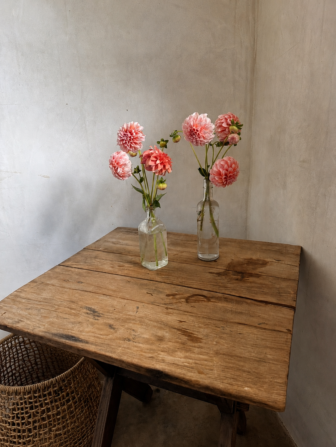 Two glass vases with pink and peach dahlias on a rustic wooden table in a corner of a room.