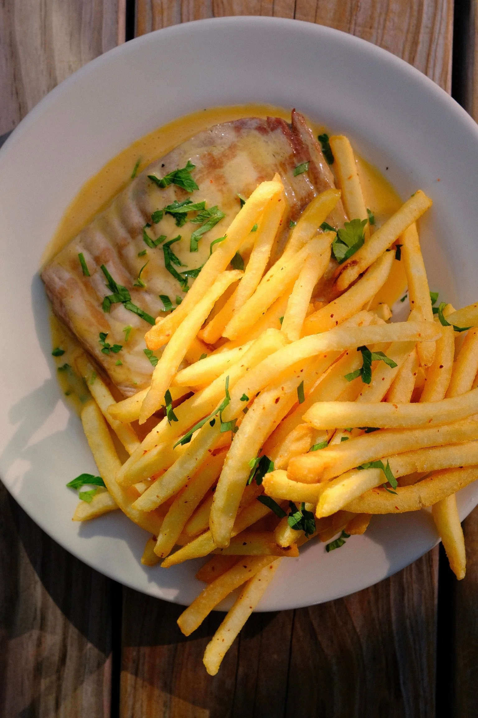 Plate of French fries topped with chopped parsley and a cheese-covered meatloaf on a wooden table.