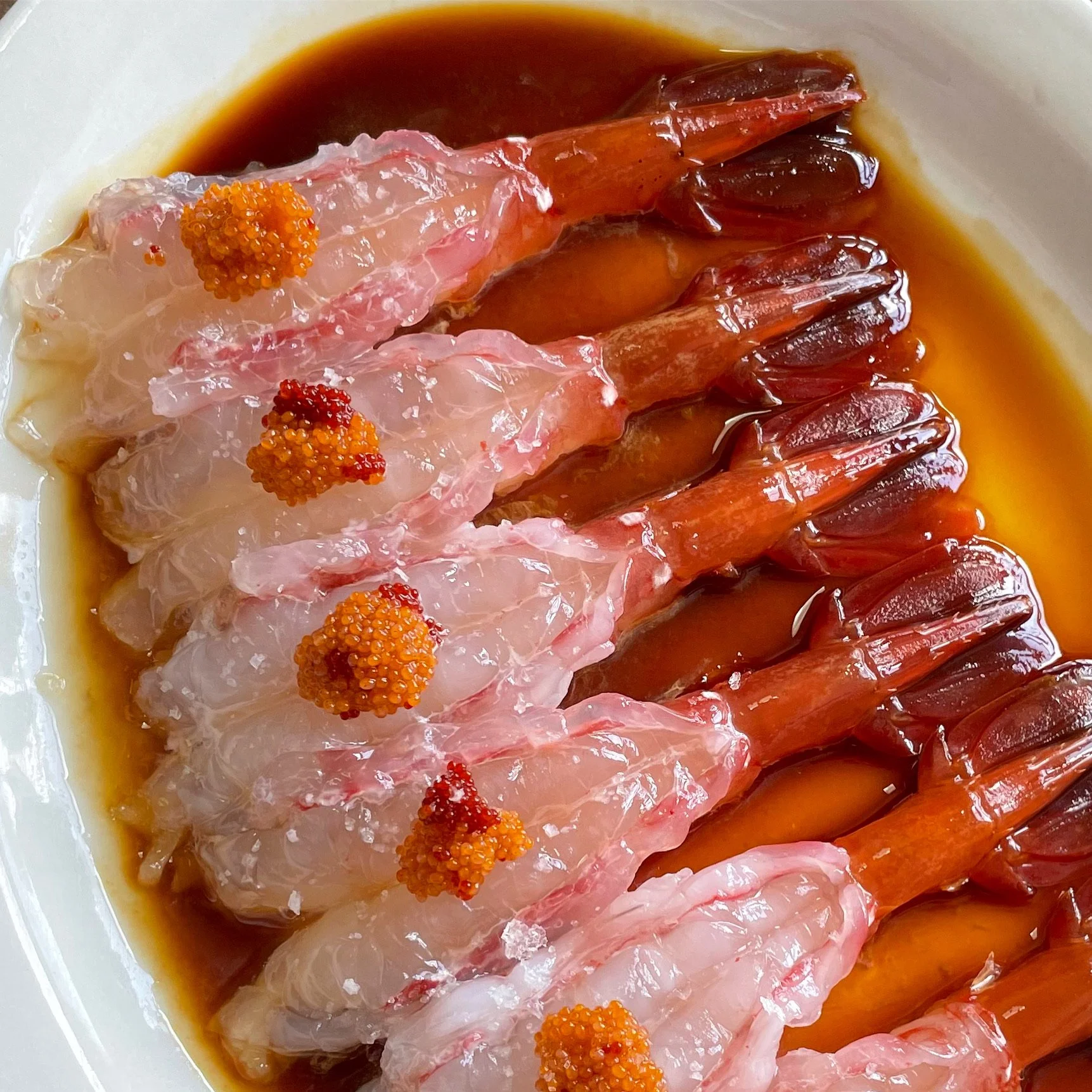 Raw shrimp topped with orange and red fish roe, served in a dark sauce on a white plate.
