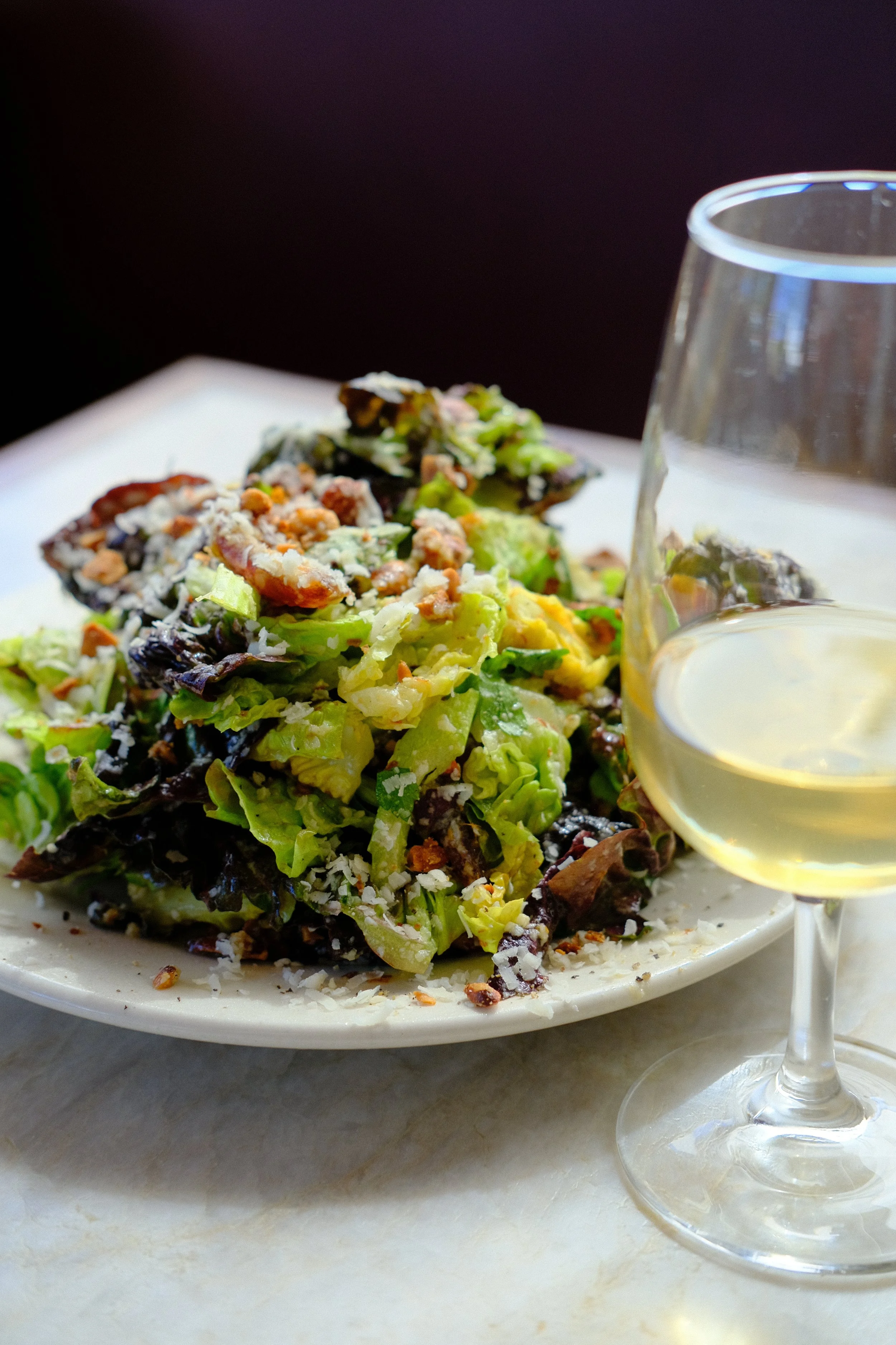 A plate of mixed greens salad with shredded cheese and nuts, accompanied by a glass of white wine on a white marble surface.