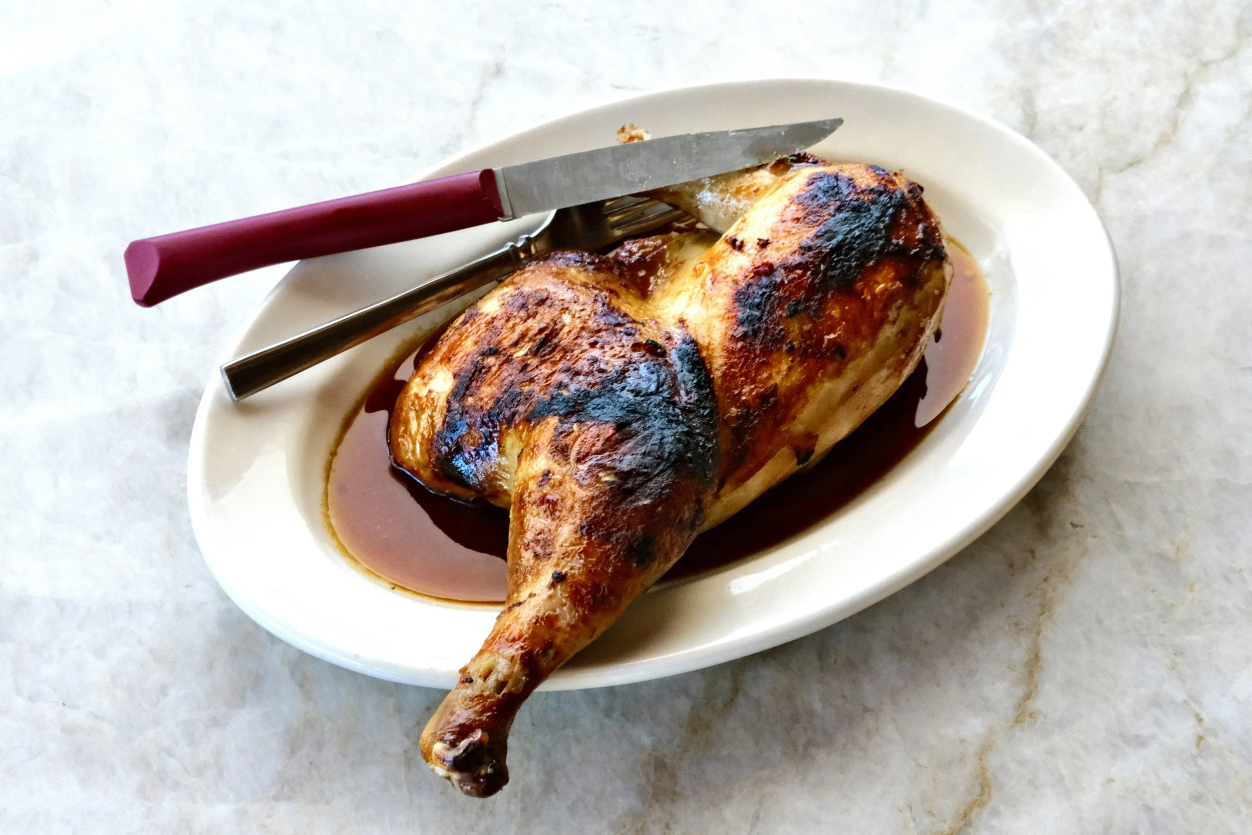 Roast chicken with crispy skin served on a white oval plate, with savory sauce, and a carving knife and fork on top.