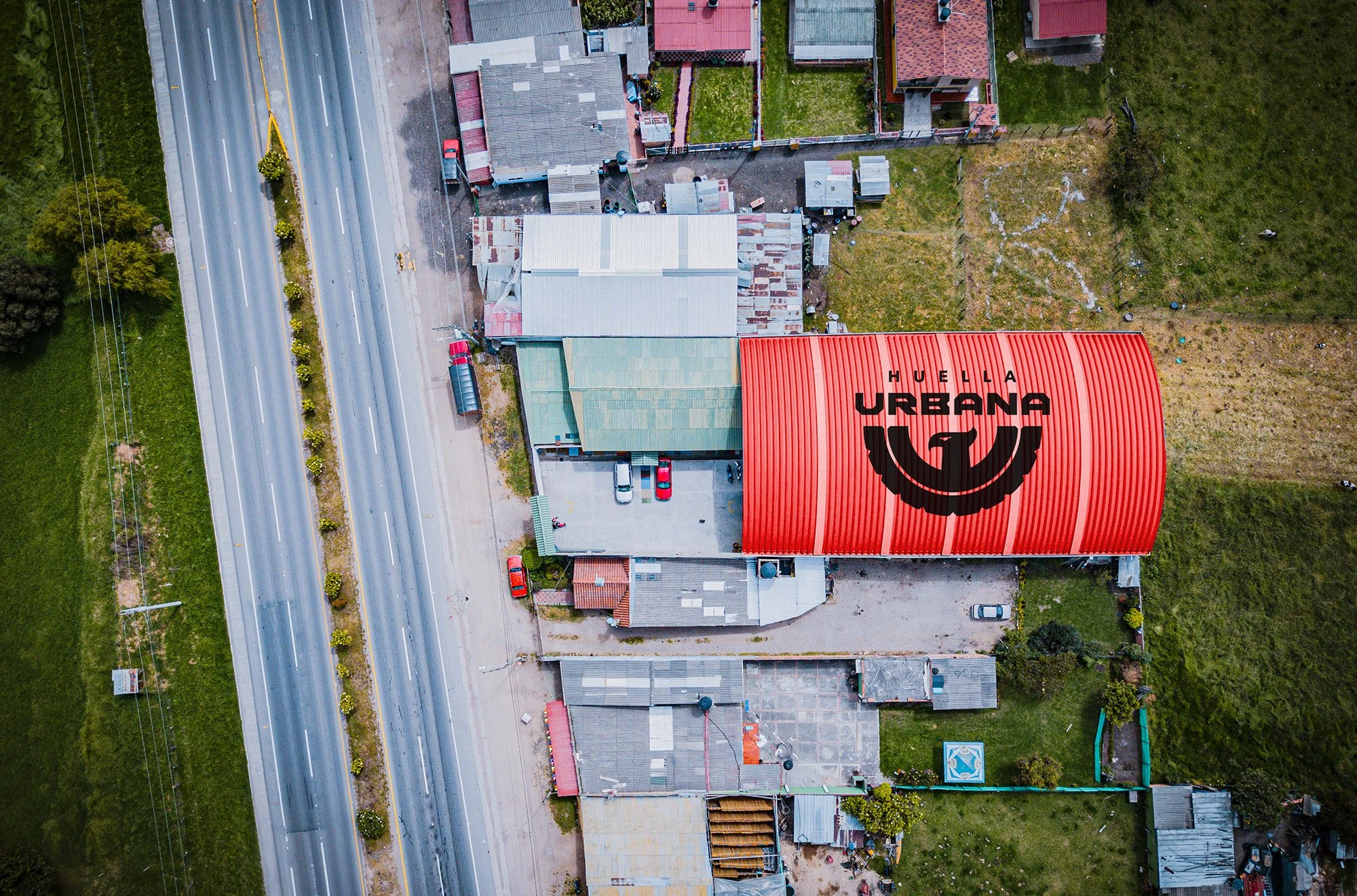 Vista aérea de un vecindario urbano con casas y un edificio con un techo rojo que tiene un logotipo y la palabra 'HUELLA URBANA'.