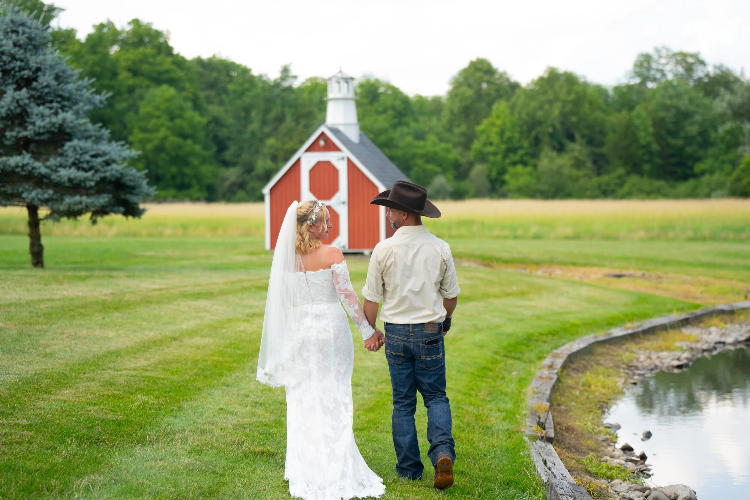 Bride and groom walking towards a red barn, surrounded by grass and trees, reflecting a rustic wedding theme.