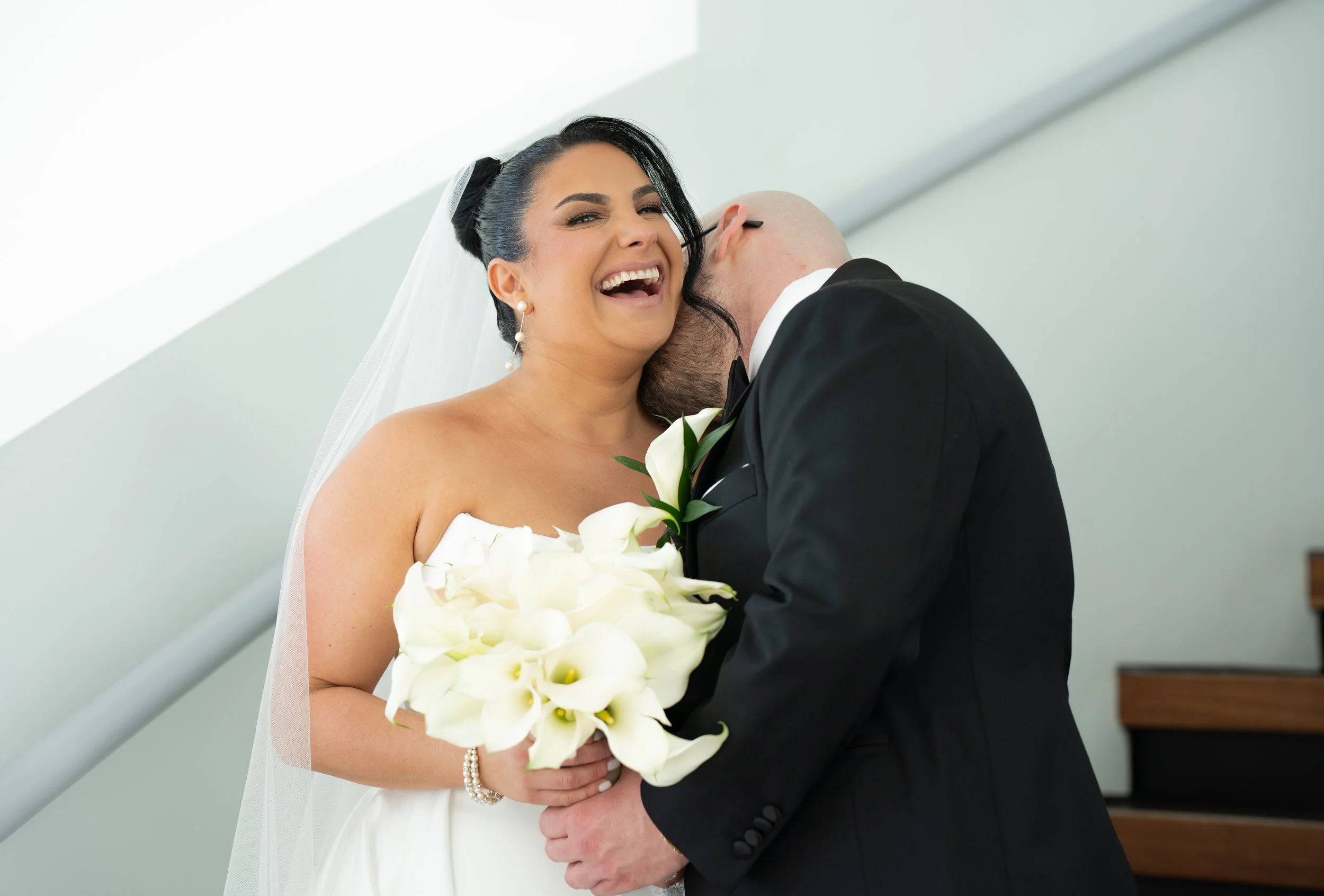 Bride and groom embracing, bride holding calla lilies, wearing white gown and veil, groom in black suit, staircase background.