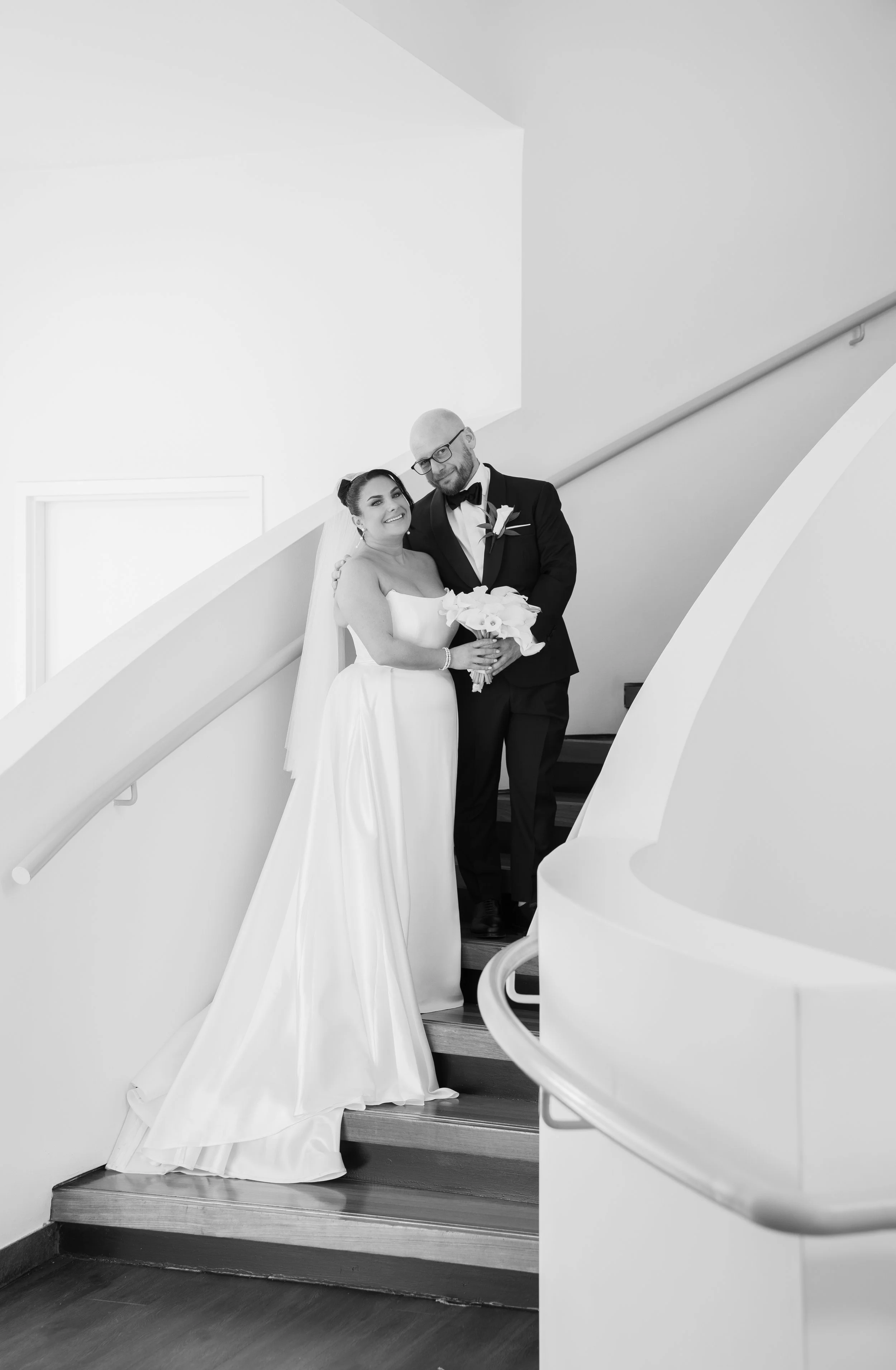 Bride and groom pose on a staircase in elegant attire; bride holds a bouquet, groom wears a tuxedo.