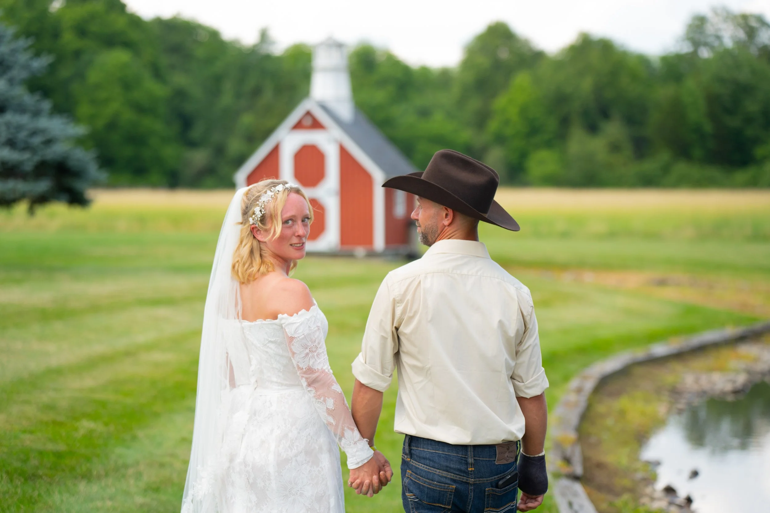 Bride and groom holding hands outdoors near a small red barn, with lush greenery in the background.