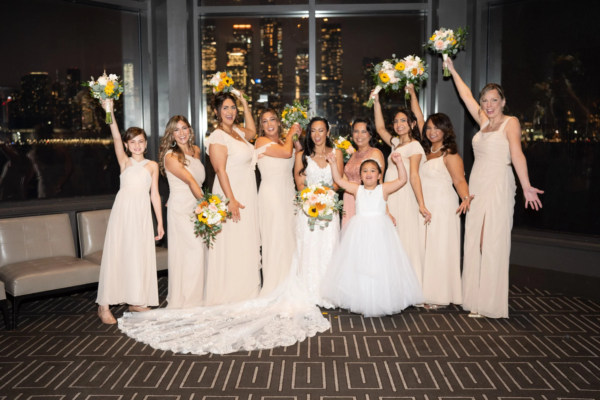 Bridal party posing indoors at night, women in light beige dresses, bride in a white gown, holding bouquets, city skyline visible through window.