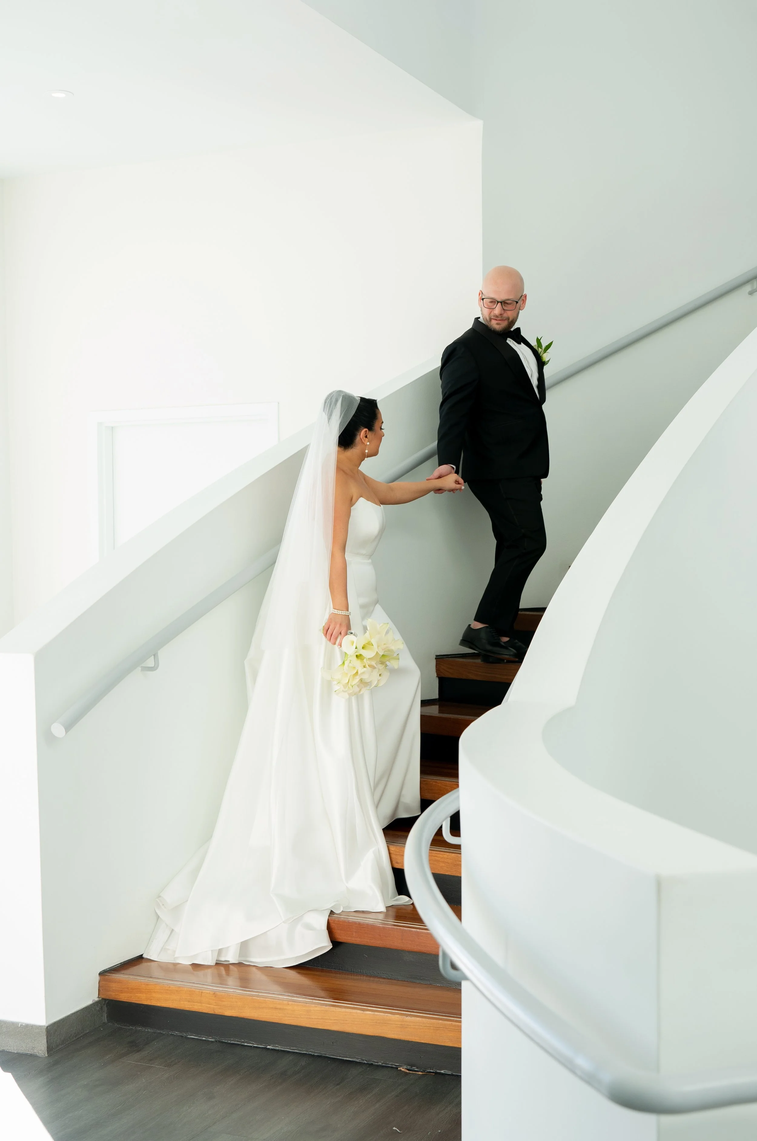 Bride and groom on staircase, bride in white gown with veil, holding bouquet, groom in black suit.